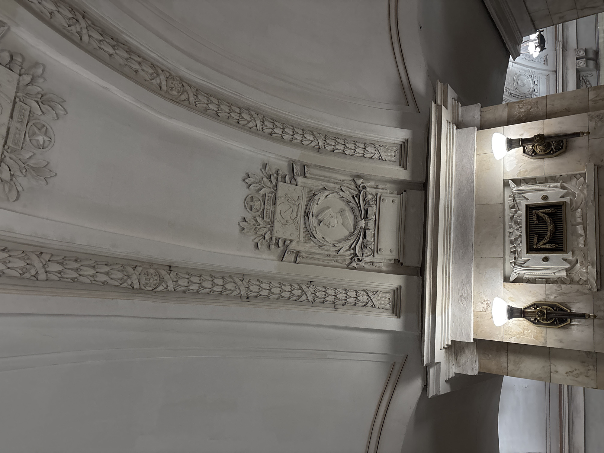 This is a low-angle, eye-level photograph of the ornate interior ceiling of a Moscow Metro station. The ceiling is white and features elaborate stucco moldings and bas-reliefs. Two arches are visible, decorated with detailed laurel wreaths and floral motifs. In the center of the upper part of the frame, a prominent Soviet-era emblem is displayed, featuring a hammer and sickle, a five-pointed star, and laurel leaves, with the inscription "EST. 1935" above it. Below this emblem, a circular bas-relief depicts the profile of a person wearing a cap. The lower part of the image shows a wall clad in light-colored marble, with two torch-like sconces providing light. One sconce is clearly visible on the left, with an ornate bronze fixture holding a glowing white light bulb. Between the sconces on the wall, there is a decorative relief panel with a grille and further ornamentation. The overall impression is one of grandeur and historical significance, characteristic of many Moscow Metro stations.