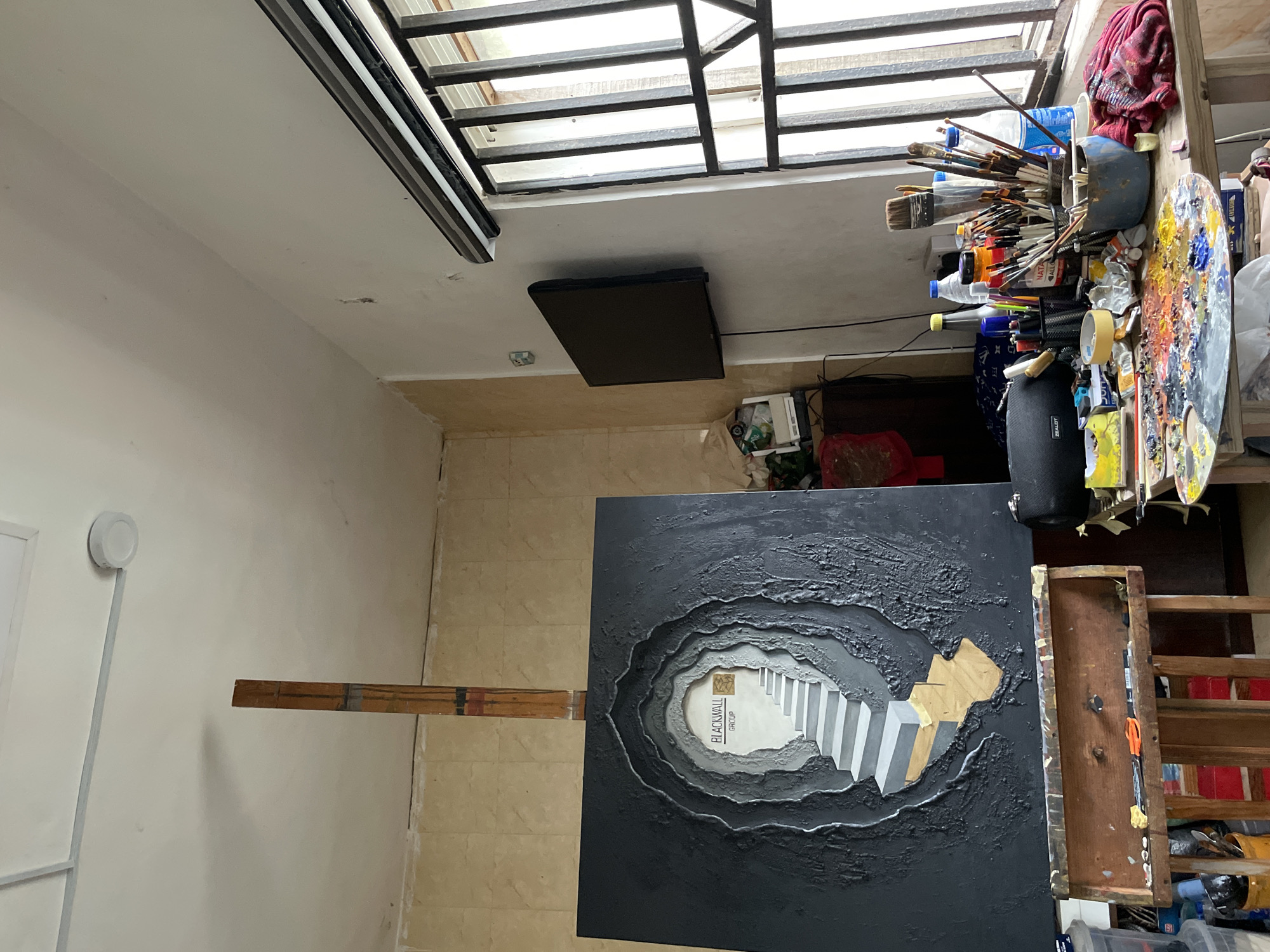 The image depicts an artist's studio in Ado, Nigeria, likely a residential or commercial space converted for creative work. The focal point is a large, textured canvas on an easel, showcasing a dark, cave-like tunnel with a brightly lit set of stairs leading upwards. The words "BLACKWALL GROUP" are visible on the wall at the end of the tunnel, above a small, square, patterned artwork. The studio environment is evident with art supplies scattered across a wooden table in the foreground. These include an artist's palette laden with vibrant paints, numerous paintbrushes of various sizes in containers, bottles of liquids, and other studio tools like scissors and tape. To the left of the canvas, another easel holds a wooden paint box with brushes and tools. In the background, a small flat-screen television is mounted on the wall, and a window with metal bars is visible on the right, allowing natural light to enter the room. The walls appear to be painted a light color, with some textured tiling on the left. The ceiling shows signs of age with some discoloration and a circular light fixture. The overall impression is of a functional, lived-in workspace, actively being used for artistic creation.