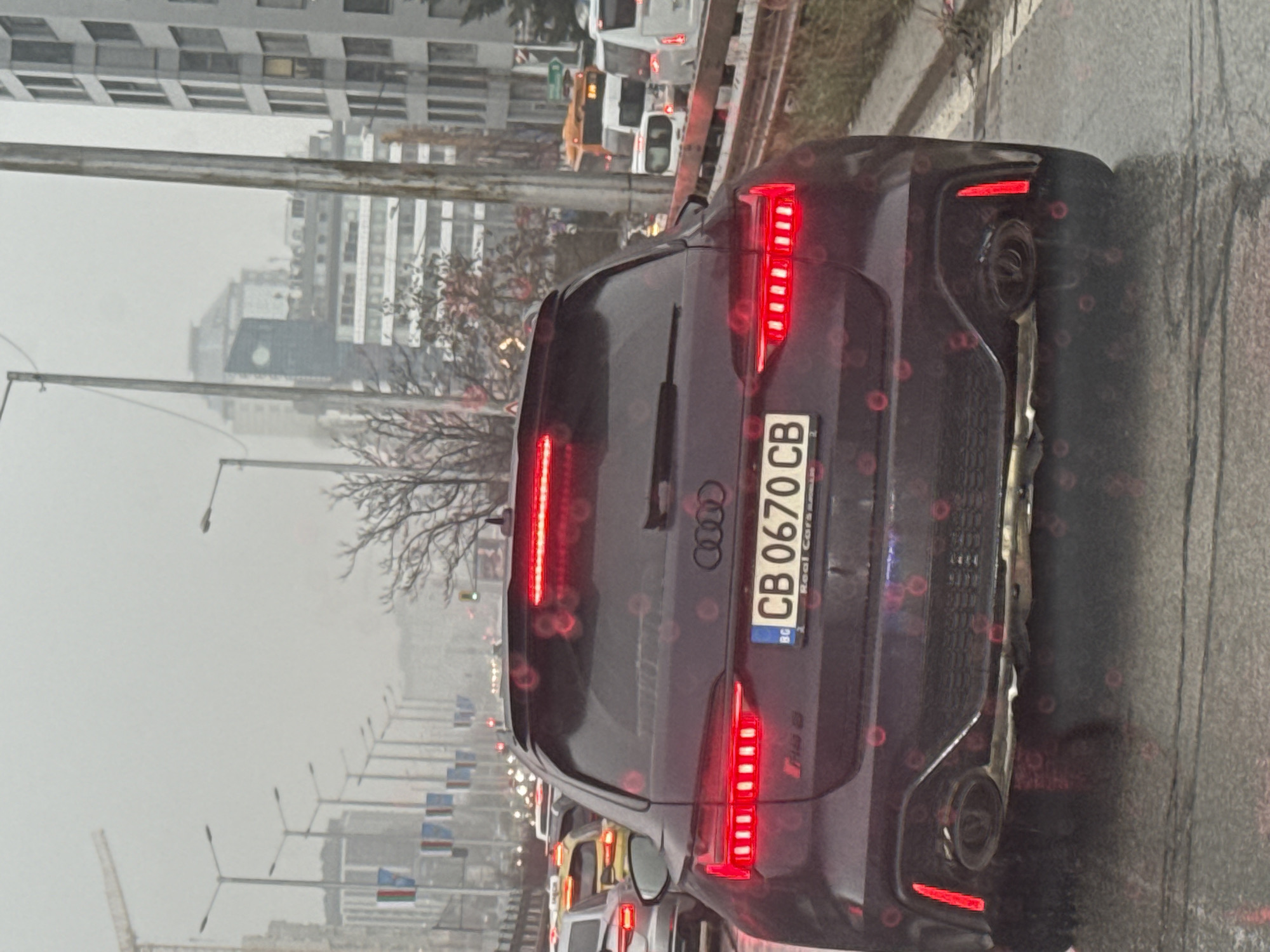 The image shows the rear of a dark gray Audi RS Q8 with Bulgarian license plate "CB 0670 CB" in Sofia, Bulgaria. The vehicle is stopped in traffic, with other cars visible ahead. The background includes buildings, street lights, and a gray, overcast sky. Raindrops are visible on the camera lens.