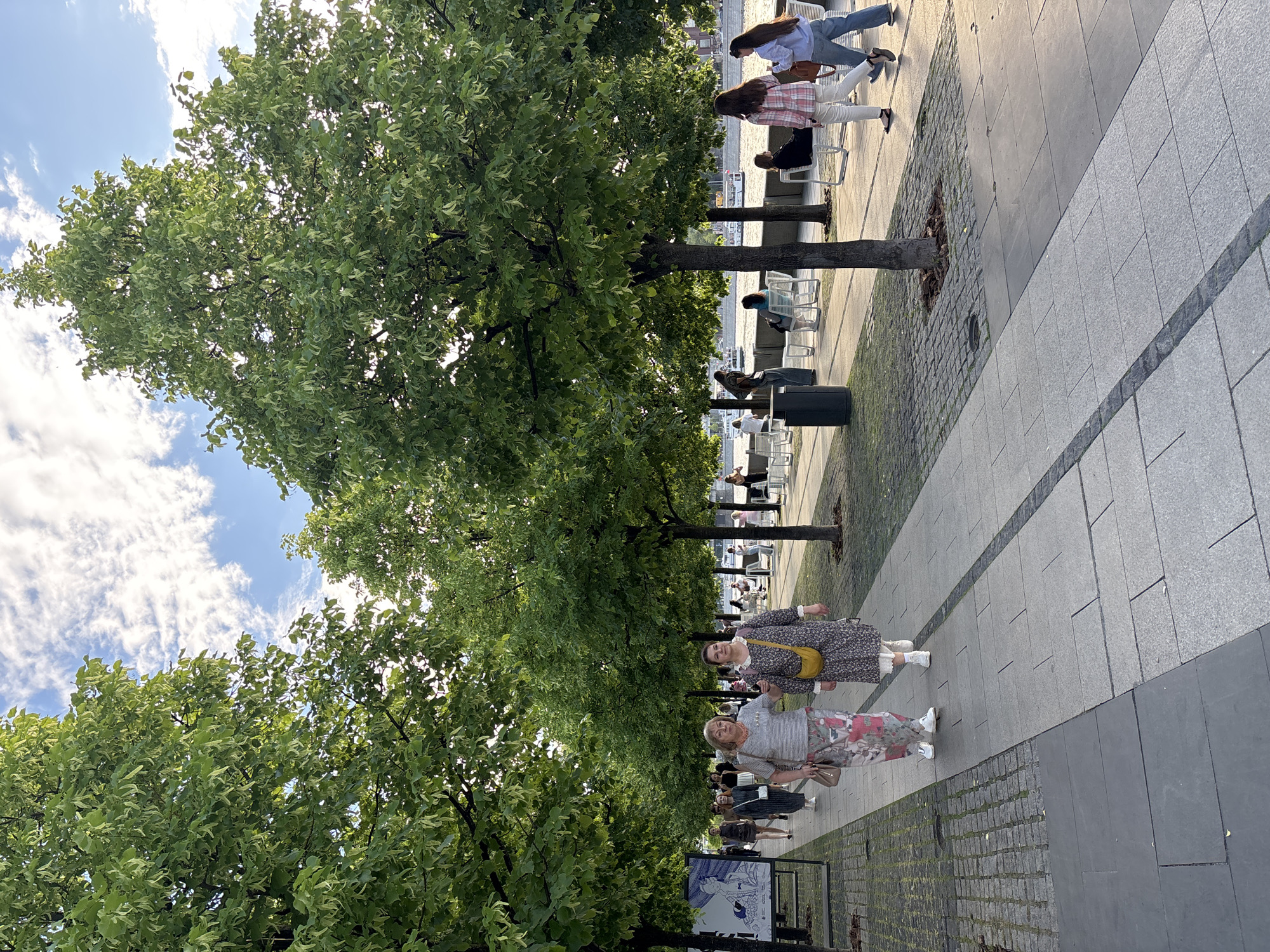 The image depicts a tree-lined pedestrian walkway in Moscow, Russia. The pathway is constructed of gray paving stones. Two women walk side-by-side toward the viewer, one in a floral print dress with a yellow purse, and the other in a striped top and floral pants, carrying a small handbag. The trees line the walkway, casting shadows on the ground. Beyond the trees are people sitting on chairs along a waterfront. The sky is blue with scattered white clouds. Street signs are visible on the left side of the path.