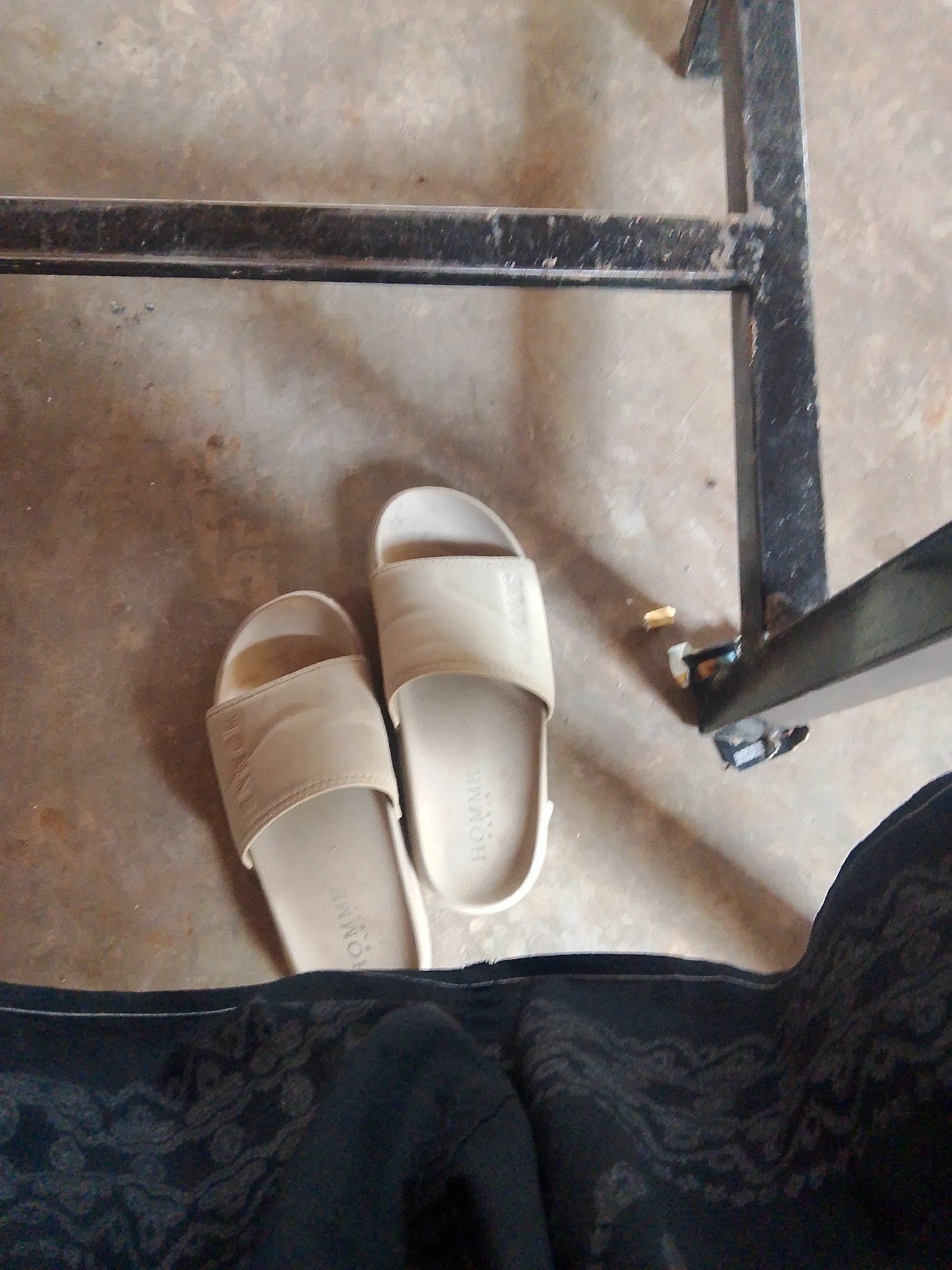 This is a high-angle shot of a pair of light beige slide sandals resting on a concrete floor. The sandals are positioned side-by-side, with the left sandal slightly behind and to the left of the right sandal. Both sandals appear to have a subtle pattern embossed on their straps, with the word "HOMME" visible on the strap of the left sandal. In the foreground, a dark fabric with a subtle pattern is visible, partially obscuring the lower portion of the sandals and the concrete floor. In the background, a black metal structure, likely part of a table or bench, is present. The concrete floor exhibits some discoloration and debris. The scene appears to be indoors or under a covered area, given the lighting and the presence of the structure. There is no identifiable indication of the specific location within Meunasahmee, Indonesia, beyond the presence of concrete and common household items.