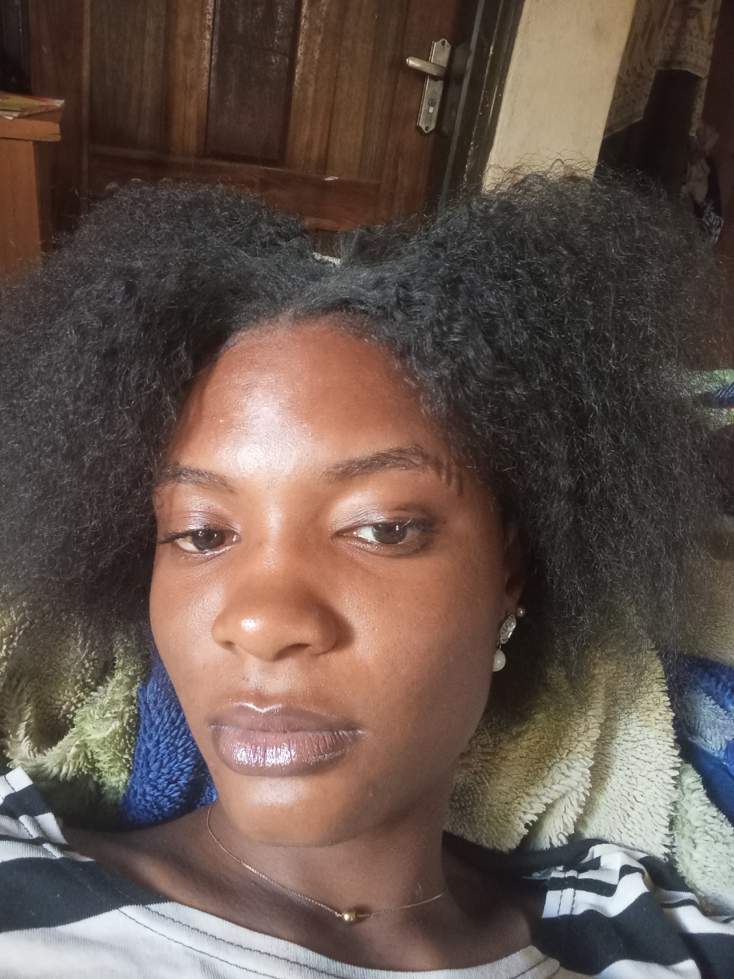 A close-up selfie of a Black woman with dark, unkempt hair. She has brown eyes, smooth skin, and glossy lips. She wears a thin gold necklace and dangling pearl earrings. She is wearing a white shirt with black stripes. A blurred background suggests she is indoors, possibly in a bedroom or living room, with a wooden door visible behind her. There is no verifiable information to conclude that this image was taken in Barikin Jirga Summa, Nigeria. The location is indeterminate.