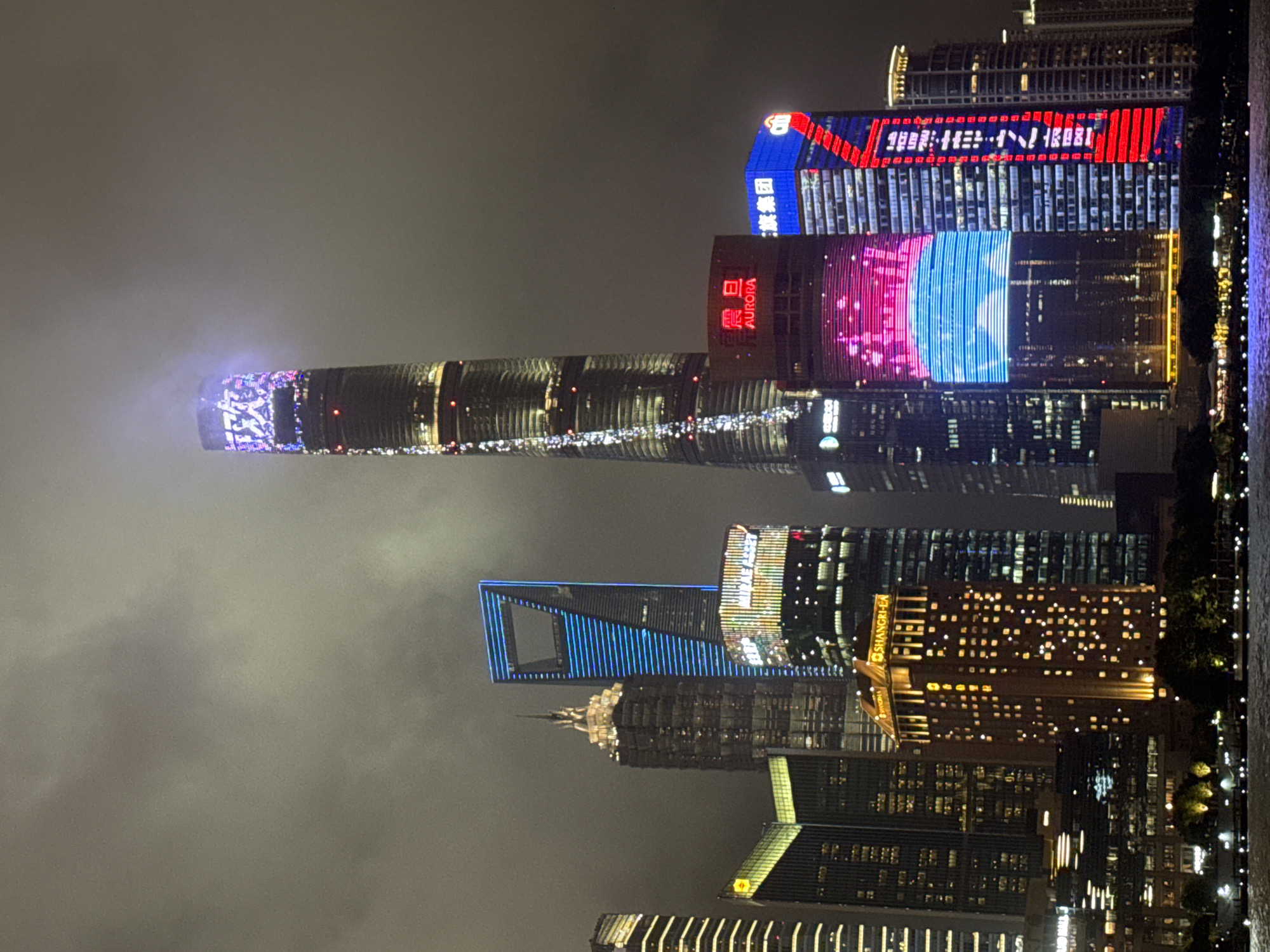 The image shows a nighttime view of the Shanghai skyline, focusing on the Pudong district. Several prominent skyscrapers are visible, illuminated by numerous lights. The tallest building in the center features a complex lighting display at its apex, with Chinese characters visible. To its right, a building displays a large digital screen showing a pink and blue animated pattern. Another building to its right is clad in blue and red lights, with visible text in Chinese characters. To the left of the tallest building, a distinctive skyscraper with a large rectangular opening at its top is illuminated in bright blue. In the foreground, a body of water reflects the city lights. Trees and a waterfront promenade are visible along the edge of the water. The sky is overcast and dark, with some haze or fog obscuring the tops of the taller structures.