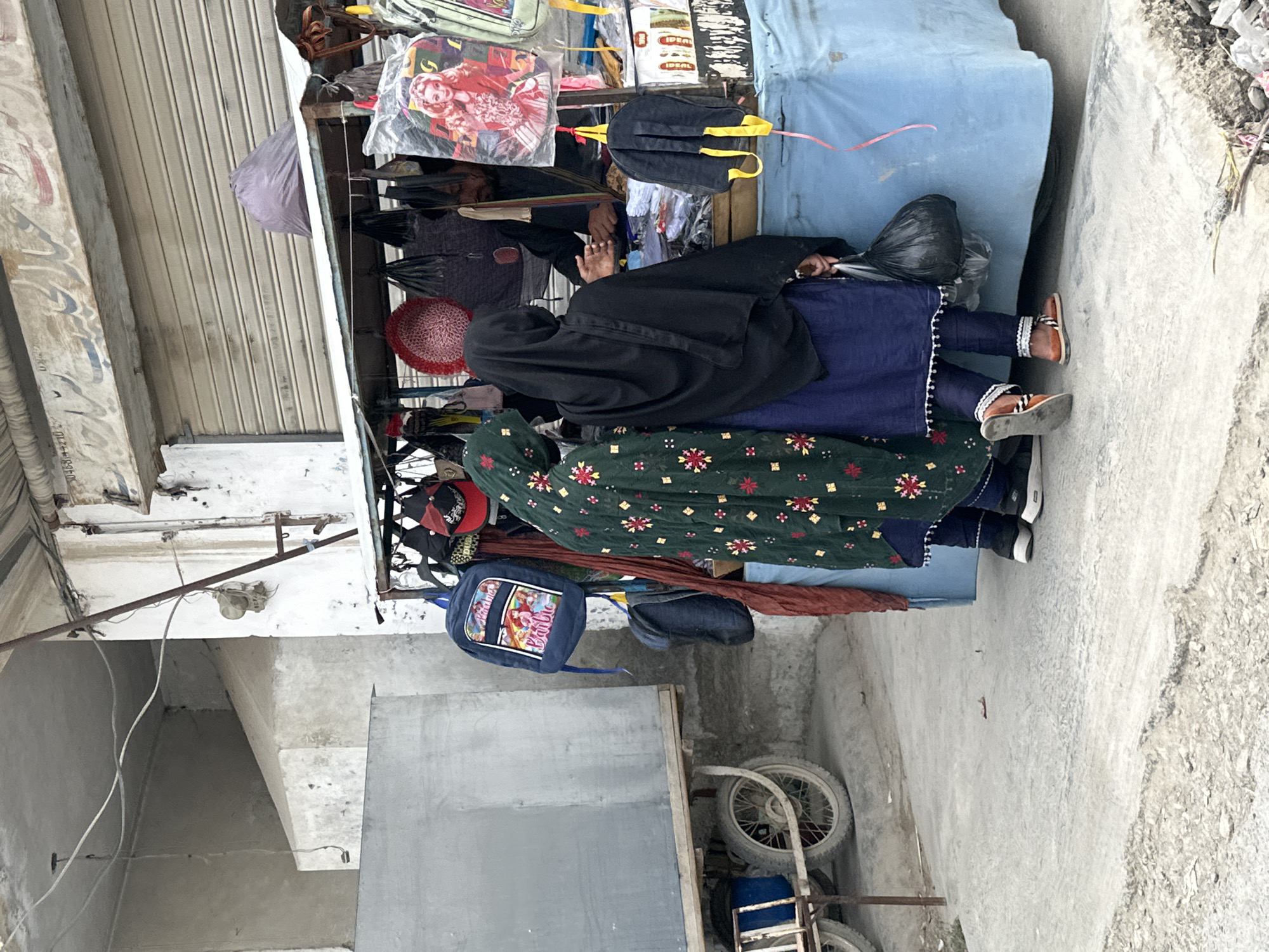 The image depicts an outdoor scene with a vendor selling merchandise from a small stand. The stand is stocked with various items including bags, backpacks, and other accessories. Two individuals, presumably customers, are visible near the stand. One is wearing a black burqa, and the other is wearing a patterned green top. The background includes architectural elements like a building with a rolling shutter door and a concrete structure. The location is an urban street, evidenced by the pavement and adjacent buildings.