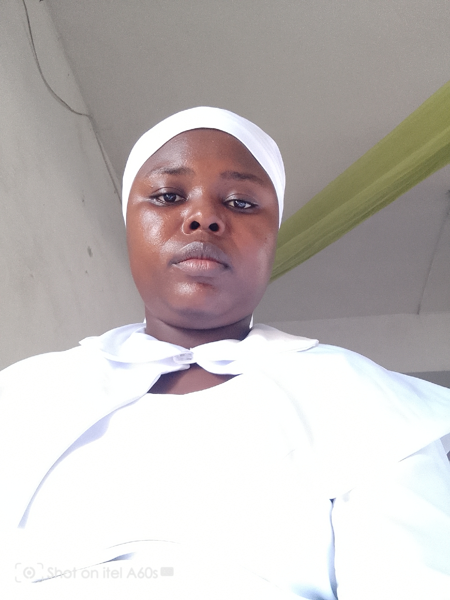 A medium shot depicts a woman with dark skin and a neutral expression, wearing a white headscarf and a white garment with a collar that ties at the neck. The background includes a pale grey wall, a visible green object suggesting a curtain or draped fabric in the upper right, and a thin electrical wire running along the upper left. The bottom of the image contains a watermark that says "Shot on itel A60s." There is no immediate confirmation regarding the exact location in Lagos, Nigeria.