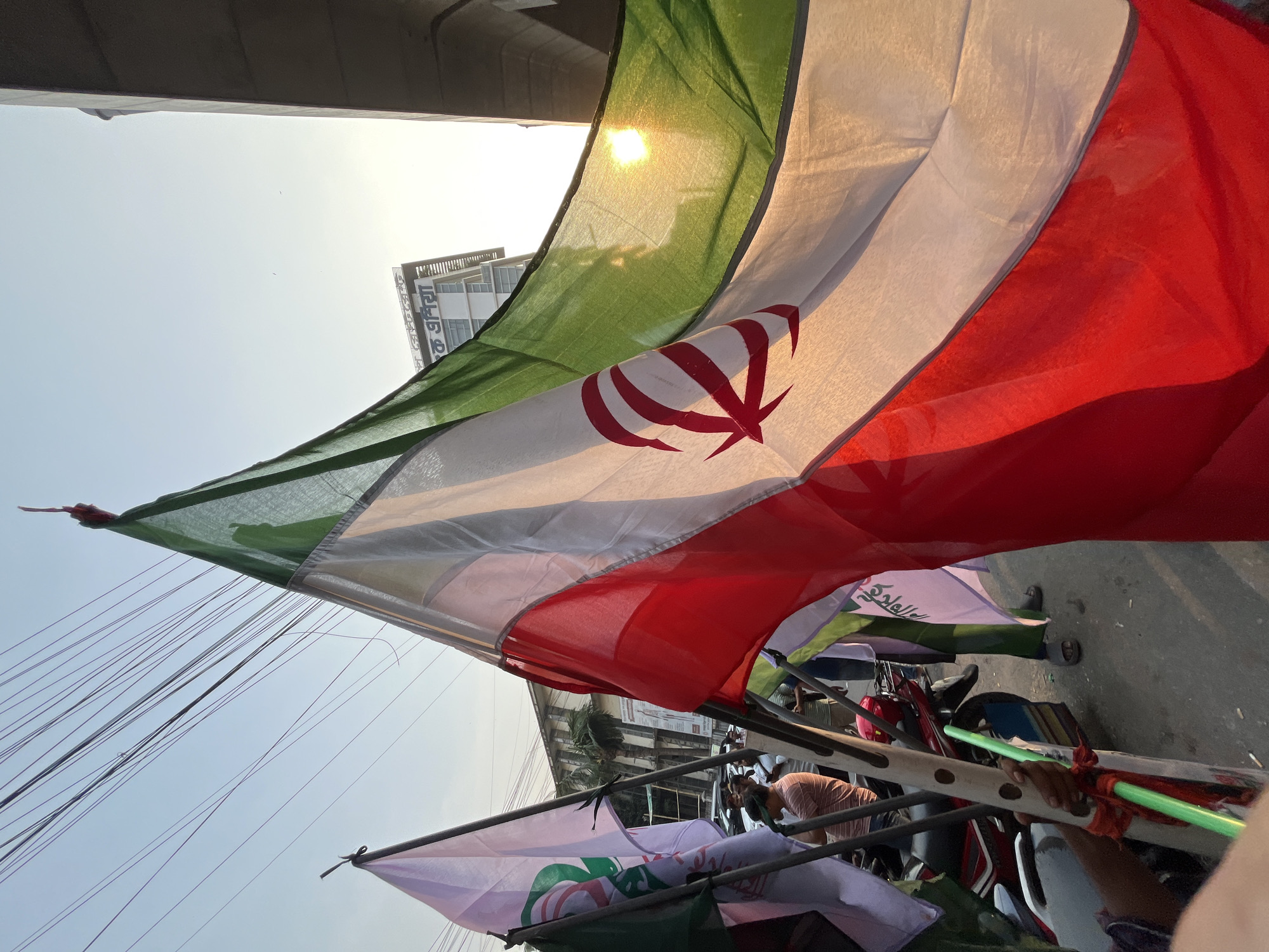 The image shows a street scene in Dhaka, Bangladesh, filled with people carrying flags. The primary flag, prominently displayed and partially obscuring the view, is green, white, and red with a stylized red emblem in the center. Below this flag, other flags with green and white colors can be seen. These are attached to poles and being held or carried by individuals. The street appears to be congested with people and motorcycles. In the background, buildings are visible under a bright, slightly hazy sky.