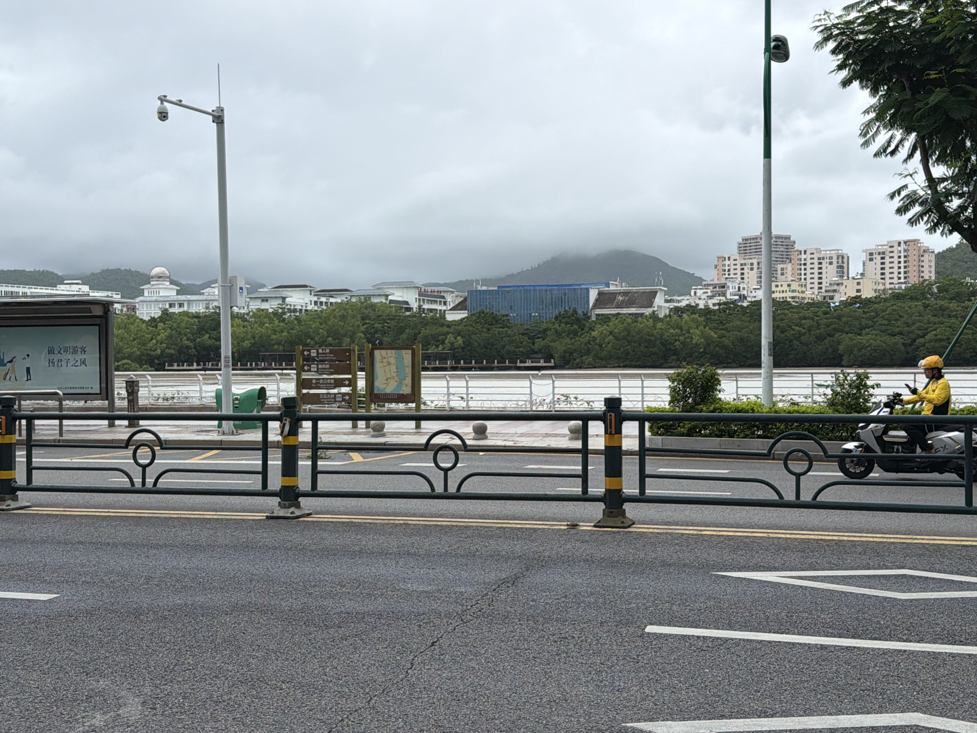 In Tianya District, China, a person wearing a yellow jacket and helmet rides a white electric scooter on a wet asphalt road. A dark green metal barrier with yellow accents separates the road from a pedestrian walkway. In the background, a body of water, lush green trees, and multi-story buildings are visible under a cloudy sky. A lamppost with a security camera is positioned near a bus stop shelter with Chinese text. The overall atmosphere suggests an overcast day in an urban coastal environment.