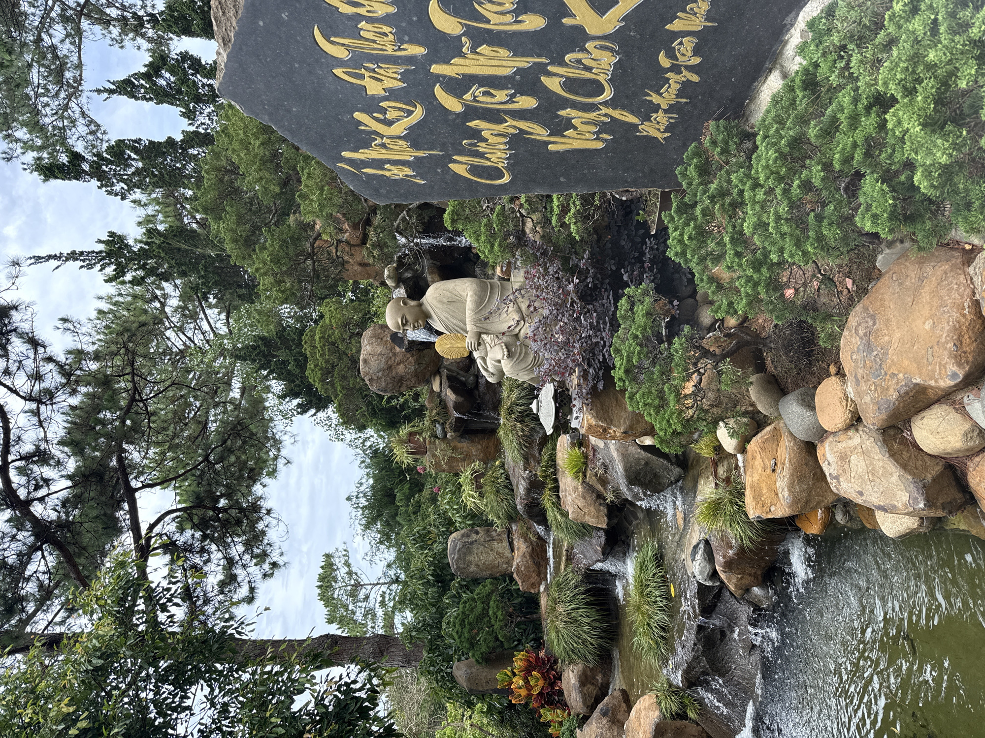 The image shows a landscaped garden in Da Lat, Vietnam. A waterfall cascades down a rocky slope into a pond. A stone statue of a seated figure with a bald head, possibly a Buddhist monk, sits in front of the waterfall. A large black stone slab with golden script stands behind the statue. The surrounding vegetation includes various trees, bushes, and grass.
