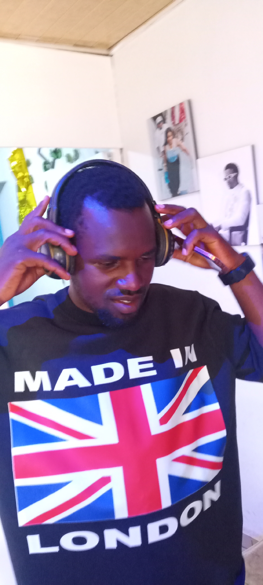 A man is indoors wearing headphones and a black T-shirt with "MADE IN" above a British flag graphic and "LONDON" below it. He is adjusting his headphones with both hands. He also has a black smartwatch on his left wrist. Two pictures are visible in the background on the wall. The lighting appears to be ambient and slightly blue-toned. The location is likely in Jos, Nigeria.