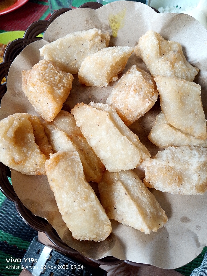 A brown woven plate holds a collection of fried, golden-brown food items. These pieces appear to be irregularly shaped, some rectangular and others more triangular or amorphous. They sit atop a sheet of tan paper, which lines the plate. Subtle textures on the fried food suggest a slightly rough or crispy surface. A few other plates and an item out of focus are in the background. A watch is visible at the very bottom.