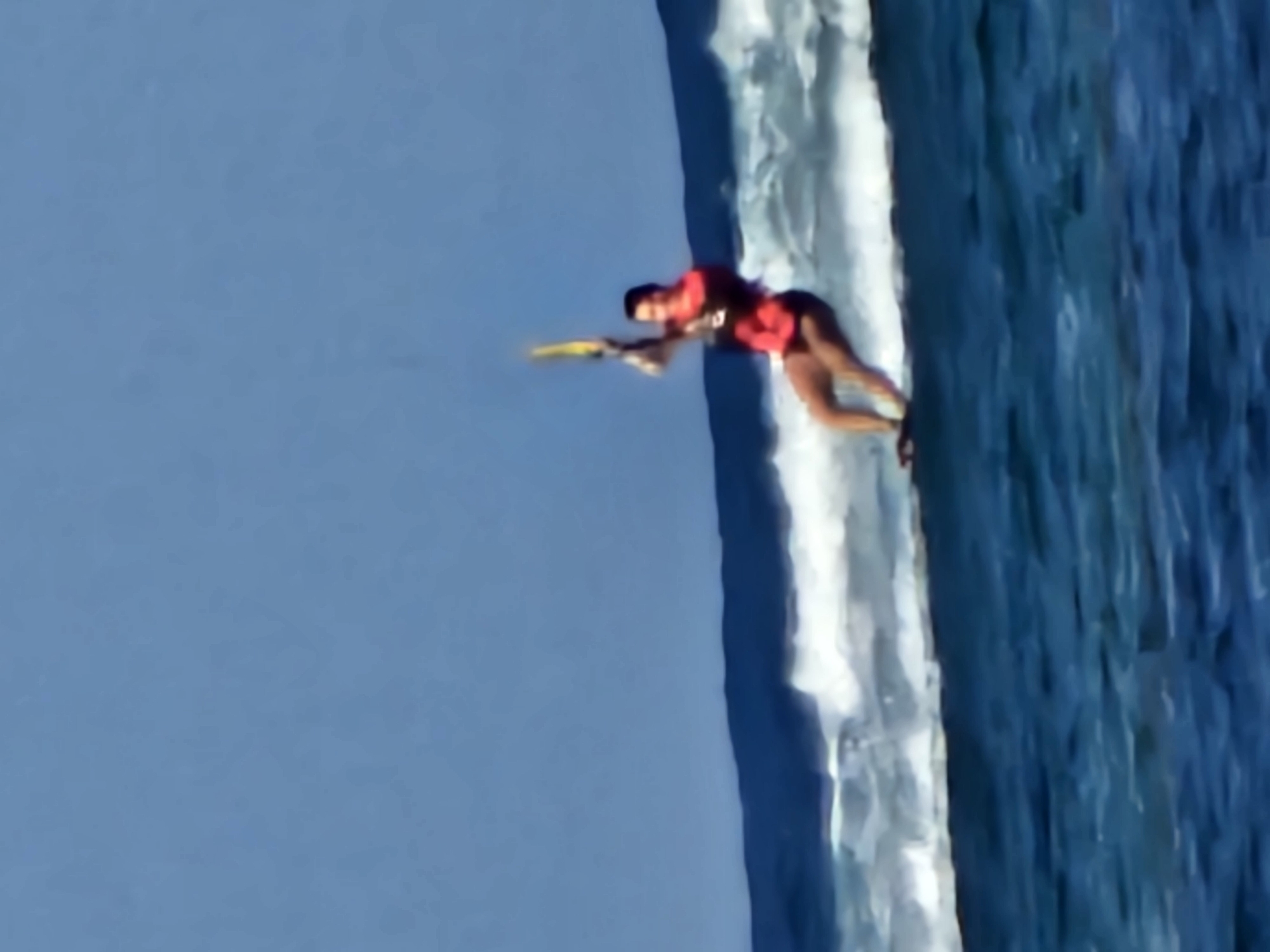 A kitesurfer in a red vest and black shorts is in mid-air, performing a jump over a wave. They are holding onto a kite bar with a yellow kite above them. The kitesurfer is on a board and is surrounded by blue ocean water with white waves crashing. The sky is clear and blue. This image is not located in Denpasar, Indonesia; it depicts a water sport.