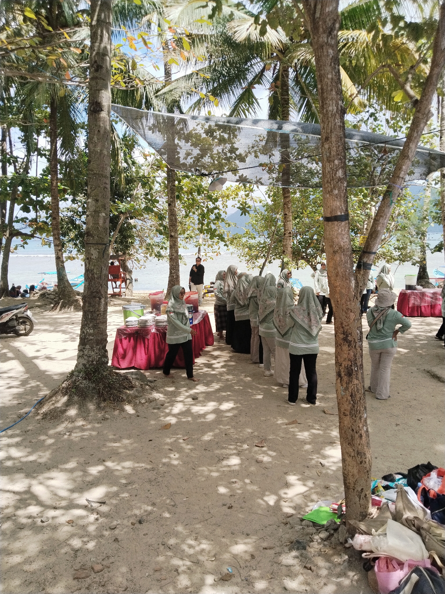 In Prigi, Indonesia, a group of individuals, mostly women in hijabs, are gathered near a beach with palm trees. They appear to be in a line near tables covered with red tablecloths, set up for some kind of event or food distribution. One person in a black shirt stands near the tables, facing the group. A motorcycle is parked to the left of the scene, and various bags are piled near a tree on the right. The environment is sandy with shadows from the trees overhead, and the ocean is visible in the background.