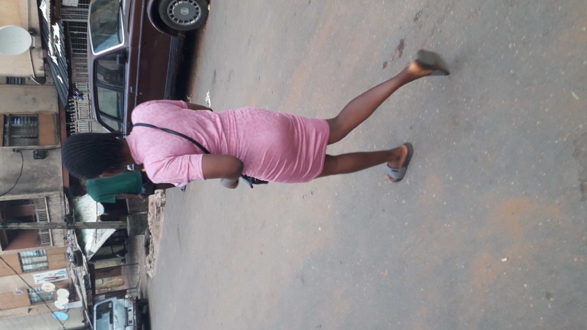 A woman in a pink dress and slippers walks on a paved street in Onitsha, Nigeria. She carries a black bag over her shoulder. A brown car is parked to her left. Buildings and a satellite dish are visible in the background, along with a truck. The street surface shows signs of wear.