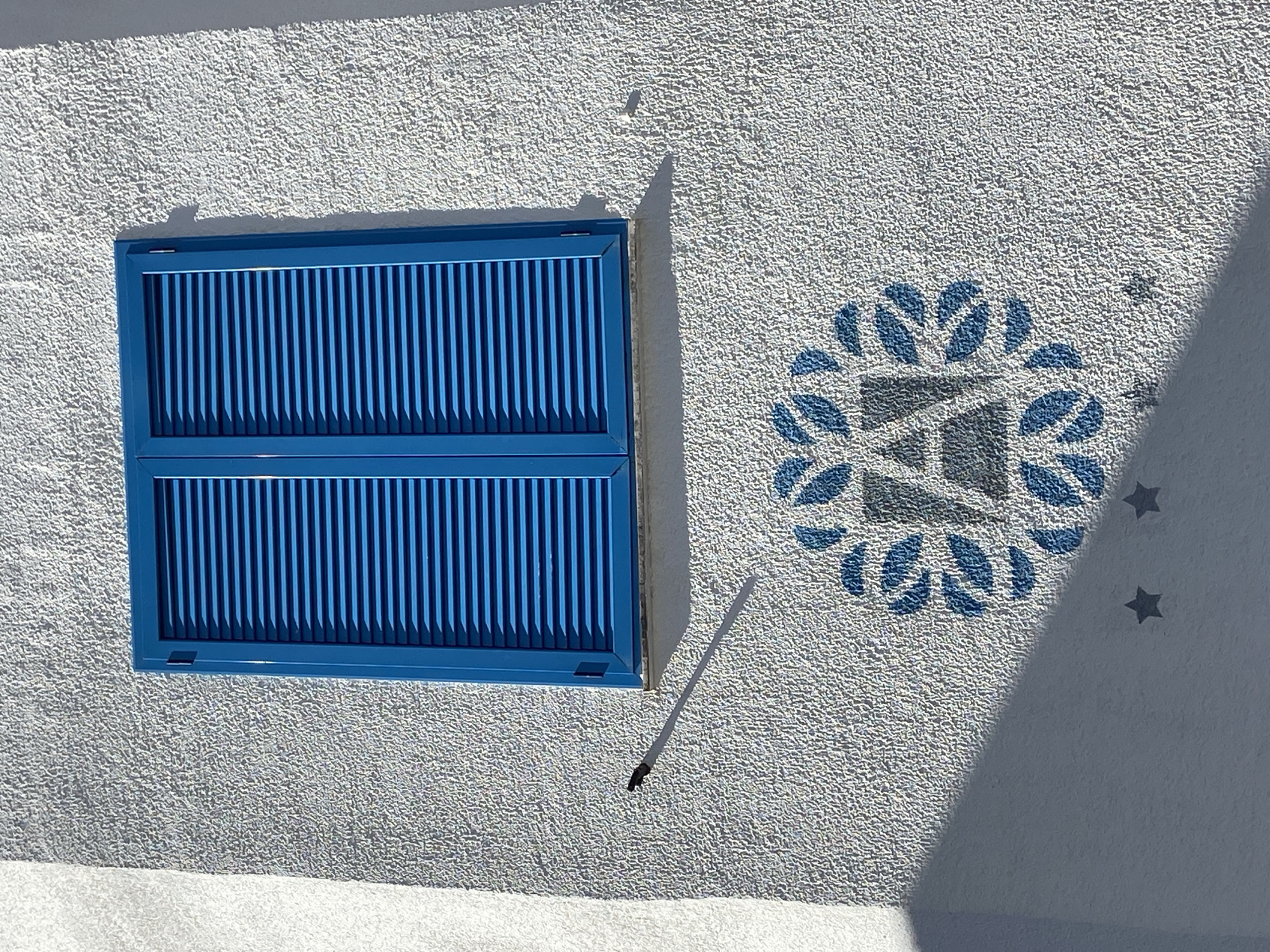 A medium shot depicts a two-paneled blue window with a slatted design, set in a white textured wall. A floral stencil pattern in blue is to the right of the window, and there are three small star stencils on the right of the pattern. A light source casts a shadow on the lower right portion of the wall. A thin, dark object protrudes from the wall, near the window. The location is Budva, Montenegro, implied by the architecture.
