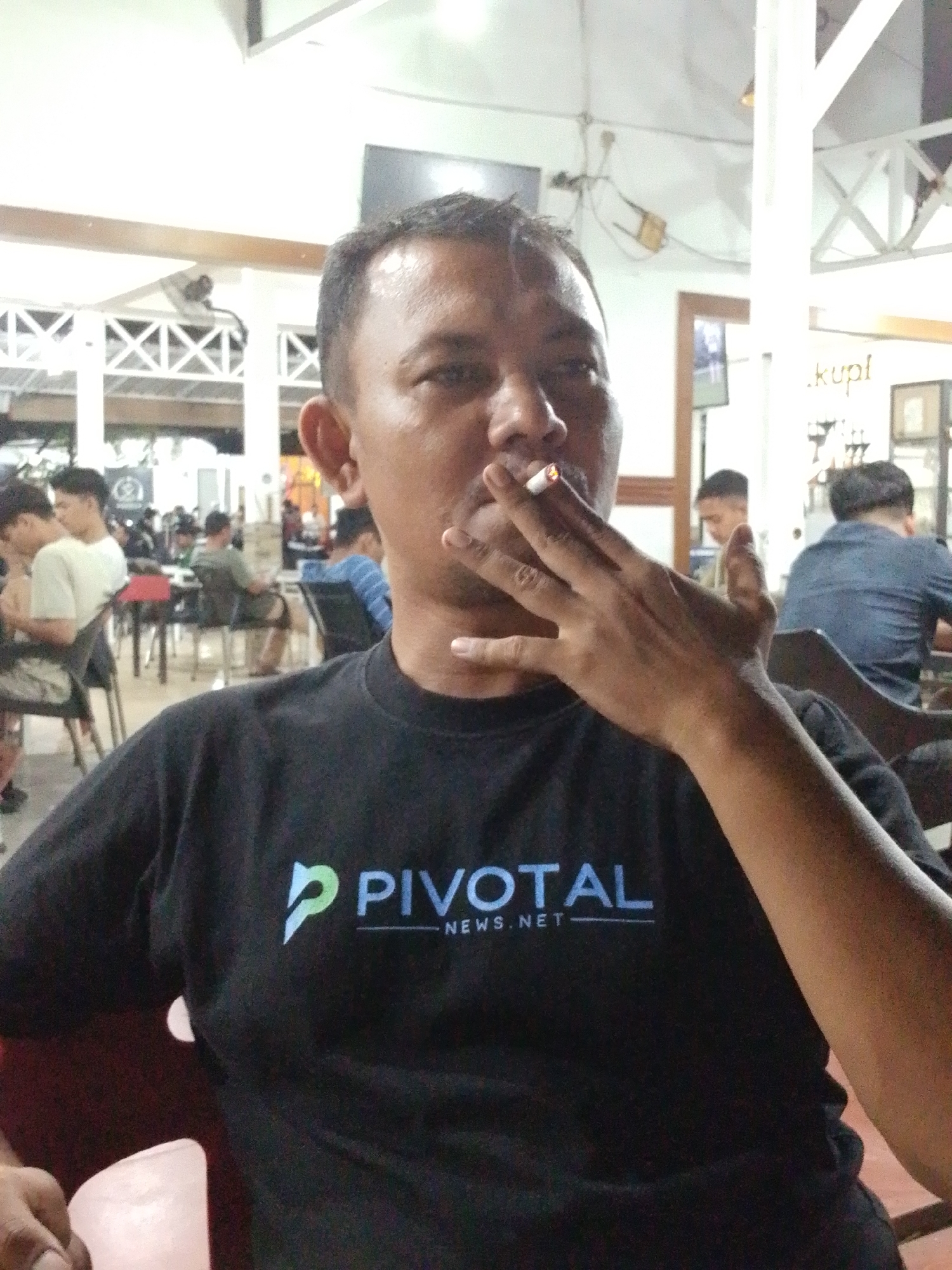 The image shows an Asian man in Lhokseumawe, Indonesia, indoors at what appears to be a cafe or restaurant. The man, wearing a black t-shirt with the word "PIVOTAL" and "NEWS.NET" printed on it, is seated and holding a lit cigarette to his mouth. Smoke is visible. Other patrons are visible in the background, seated at tables. The interior has a casual setting with ceiling fans and open views of the surroundings.