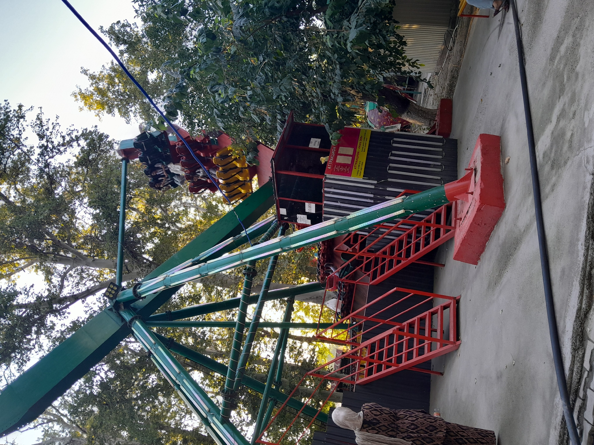An amusement park ride, commonly known as a pendulum ride or swing ride, is depicted in an outdoor setting. The ride features a large green arm structure supporting a rotating gondola at the end, currently occupied by several passengers. Red metal staircases lead up to the ride platform. A person wearing a headscarf and a patterned dress stands to the left, facing away from the ride. The ground is paved with concrete, and mature trees with lush green foliage surround the area. A black hose or cable runs across the foreground. The scene appears to be in the city of Fergana, Uzbekistan, based on the presence of Cyrillic script on signage, which is characteristic of the region.