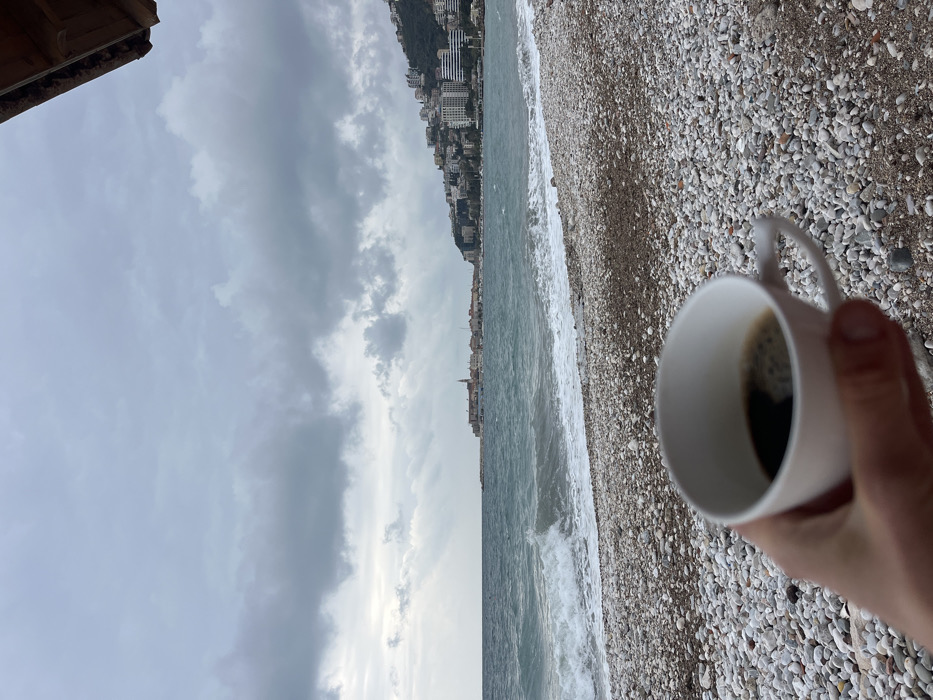 The image displays a coastal scene in Budva, Montenegro, captured from a low vantage point on a pebble beach under an overcast sky. In the immediate foreground, a human hand holds a white ceramic cup containing dark liquid, likely coffee, with a layer of crema visible on the surface. The hand and cup are slightly out of focus. The midground features a beach composed of various-sized light grey and white pebbles, transitioning into the turquoise-green sea. Moderate waves are visible breaking onto the shore, creating white foam. In the distant background, the historic Old Town of Budva is visible to the left, characterized by its cluster of red-roofed buildings and a prominent church steeple, extending into the water via a stone breakwater. To the right of the Old Town, the modern urban landscape of Budva ascends a verdant hillside, showcasing numerous multi-story residential and commercial buildings. The sky above is heavily clouded with dark grey formations, though a lighter band is present near the horizon line. A segment of a brown wooden architectural element is visible in the upper right corner, suggesting an overhead structure.