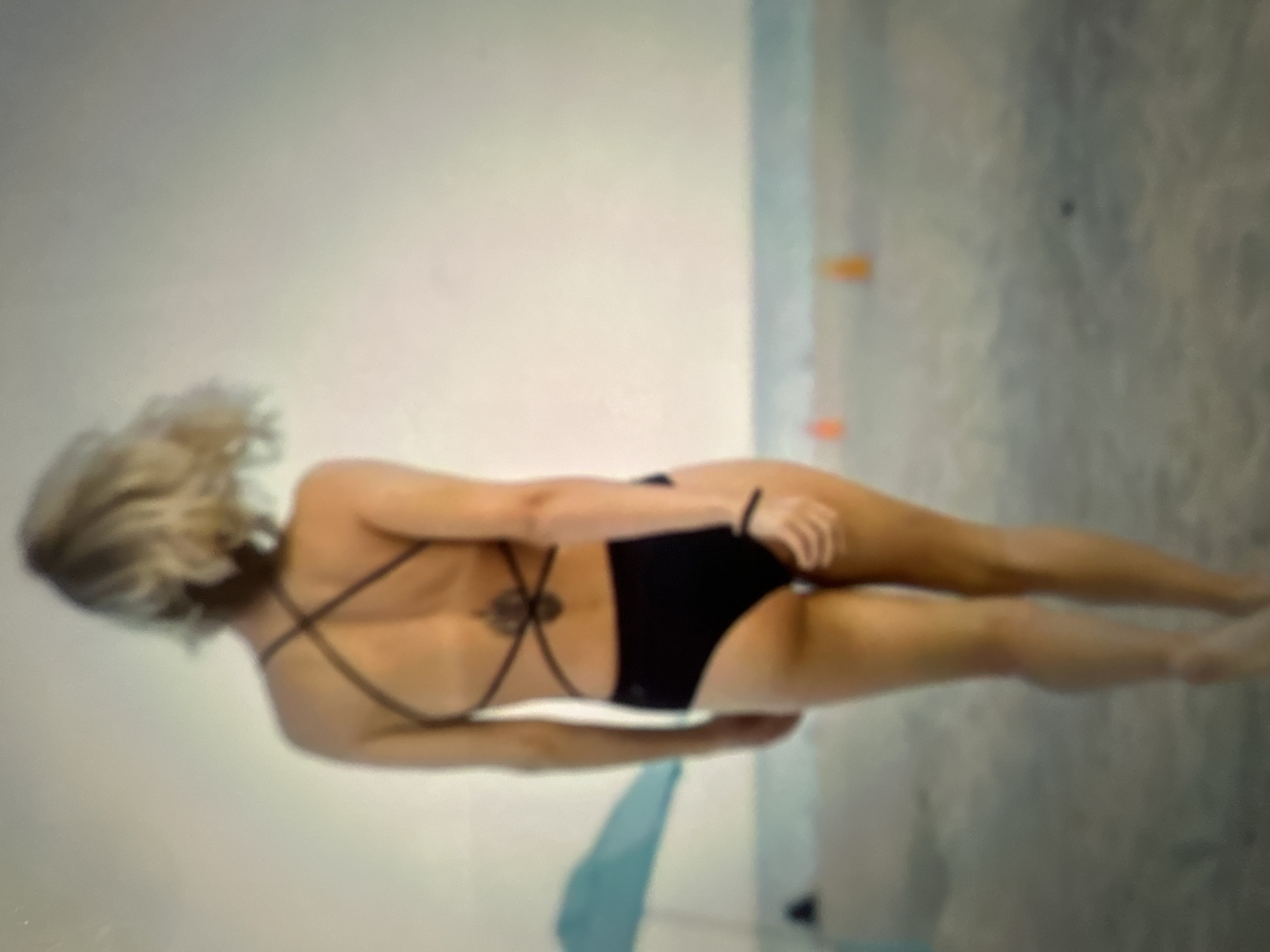 The image depicts a person walking away from the camera on a sandy beach. The person is wearing a black swimsuit and has short, blonde hair. Their back is visible, and there is a tattoo on their lower back. In the background, there is a light blue beach umbrella and two orange cones. The sky is overcast, and the sea is visible in the distance. The image quality is blurry, suggesting it may have been taken with a low-resolution camera or from a distance. The location is not identifiable as Gelendzhik, Russia, or any specific city.