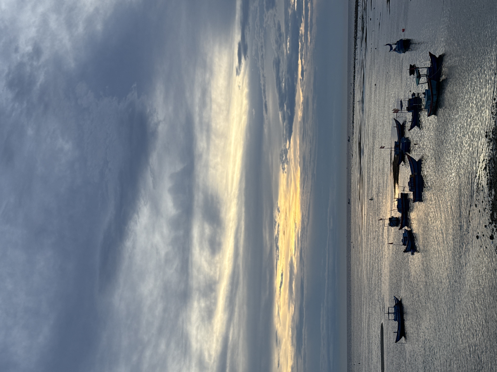 A wide shot captures a serene seascape at dusk, showcasing a tranquil sea with several small fishing boats anchored near the shore. The sky above is a canvas of dramatic clouds, with streaks of warm yellow and orange from the setting sun breaking through the diffused gray tones. The water reflects the sky's muted colors, creating a shimmering effect. In the foreground, a rocky shoreline leads into the shallow water where the boats are moored. The boats, silhouetted against the water, are diverse in size and design, some with flags, suggesting a fishing community. The overall atmosphere is peaceful and contemplative, hinting at the end of a day's work at sea. This scene is likely located along the coast of Lý Sơn Island, known for its fishing villages and beautiful twilight skies.
