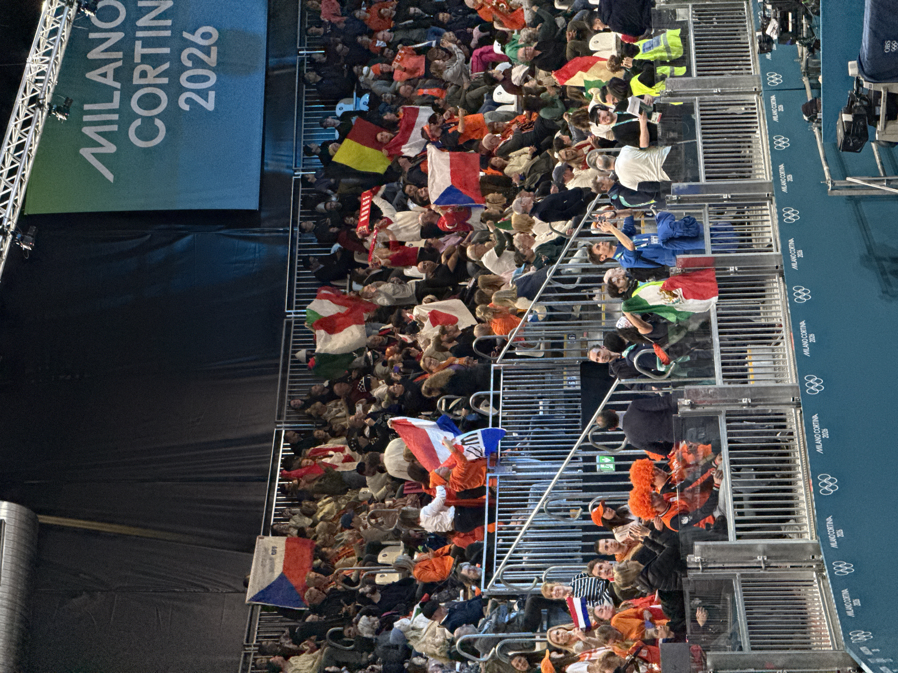The image captures a bustling scene from an indoor or covered event, showcasing a large crowd of spectators and official branding for the Milano Cortina 2026 Olympic and Paralympic Games.
In the upper left portion of the image, a prominent banner is displayed, featuring the white text "MILANO CORTINA 2026" on a blue-green background. A metal truss structure, commonly used for staging or lighting, is visible to the immediate left of this banner.
The main body of the image is filled with a dense crowd of people seated in tiered sections. Many spectators are actively holding up national flags, indicating an international gathering. Clearly visible flags include:
* A Hungarian flag (red, white, and green horizontal stripes).
* A Belgian flag (black, yellow, and red vertical stripes).
* A Japanese flag (a red circle on a white background).
* A Czech Republic flag (white and red horizontal stripes with a blue triangle on the hoist side).
* A French flag (blue, white, and red vertical stripes).
* A Mexican flag (green, white, and red vertical stripes with an emblem in the center).
Several individuals in the crowd are also seen wearing distinctive bright orange clothing, including orange wigs, strongly suggesting support for the Netherlands. The spectators are dressed in a variety of casual attire, with some wearing jackets, possibly indicating cooler temperatures or an evening event. They appear engaged, watching an unseen event, with some individuals holding phones or taking photos.
To the right side of the crowd, a series of metal barriers separates the spectator area from what appears to be an official zone. Along this barrier, a continuous blue banner displays the "MILANO CORTINA 2026" logo alongside the Olympic rings symbol, repeated multiple times. In this area, several individuals are present, including one person wearing a bright yellow safety vest, likely event staff. Professional camera equipment, including a large camera on a stand, is visible, suggesting media coverage. Another person is seen wearing a blue uniform with "OBS" (likely Olympic Broadcasting Services) visible on their attire.
The overall setting appears to be a stadium or arena, illuminated by artificial lighting. The presence of numerous national flags and the "MILANO CORTINA 2026" branding strongly suggest this is an event related to the upcoming Winter Olympics and Paralympics, possibly an opening ceremony, a sporting competition, or a celebratory gathering.