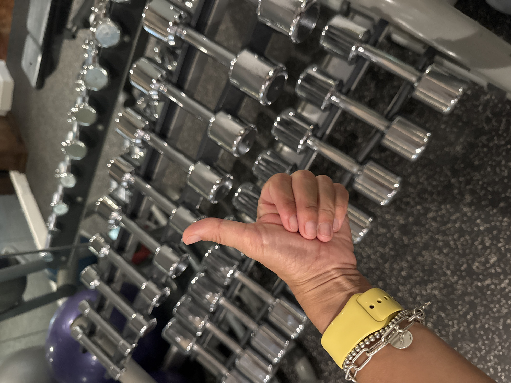 The image shows a hand giving a thumbs-up in front of a rack of dumbbells. The hand is wearing a yellow watch band and a silver bracelet. The dumbbells are arranged in rows on a metal rack. The background is a dark, possibly textured surface. The location is likely a gym, but the city of Kuta Selatan, Indonesia, is not discernible from the visual information provided.
