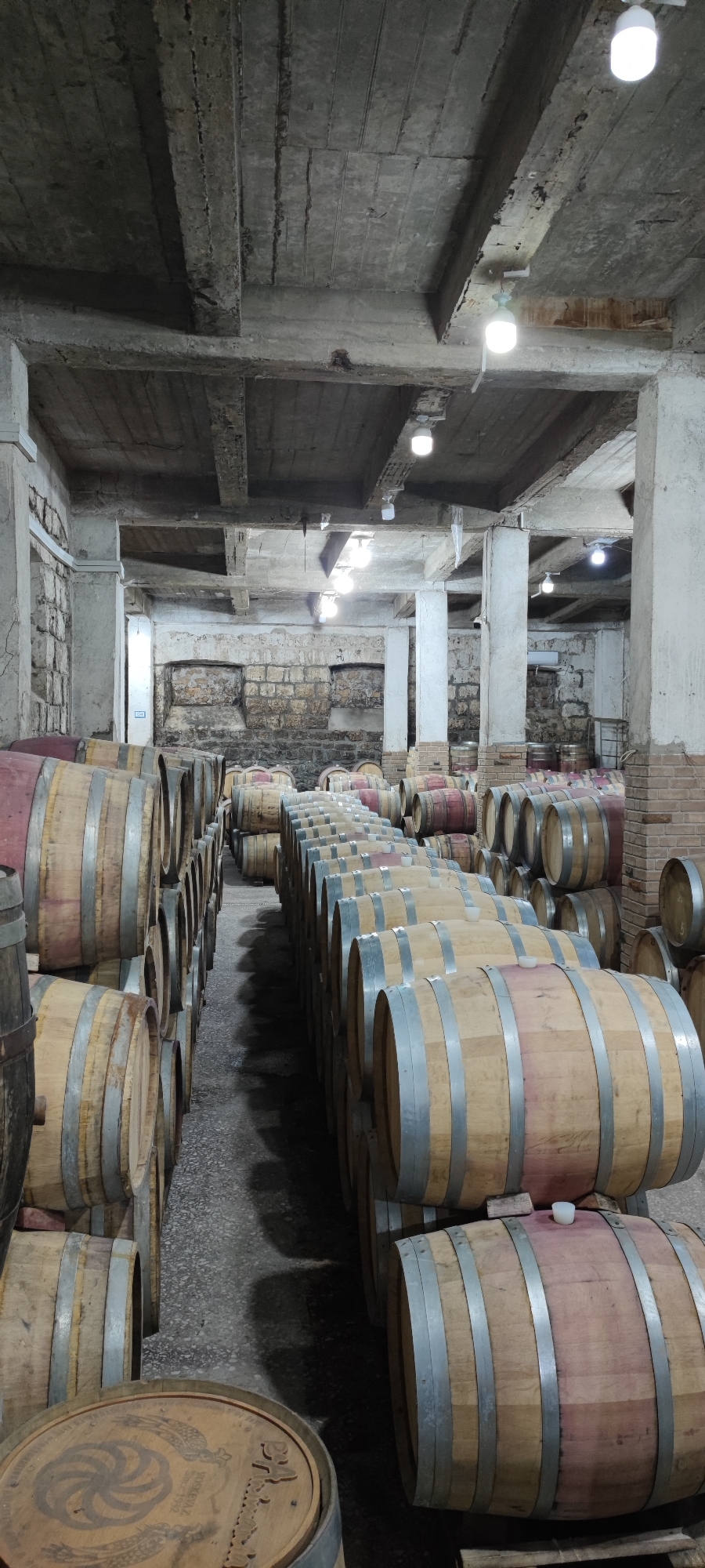The image depicts a dimly lit interior of a winery or cellar, likely located in Voskevaz, Armenia. Rows upon rows of wooden barrels, some with reddish-brown tops and others with natural wood, are meticulously arranged. The barrels vary in size and are stacked in some areas, suggesting a storage facility for wine or spirits. The architecture of the room is rustic, with exposed concrete ceilings supported by robust columns, and walls constructed from stone and brick. Several white lights are suspended from the ceiling, illuminating the scene with a practical but somewhat stark light. The overall atmosphere is one of aged tradition and industrial production within the beverage industry. No individuals are present in the frame.