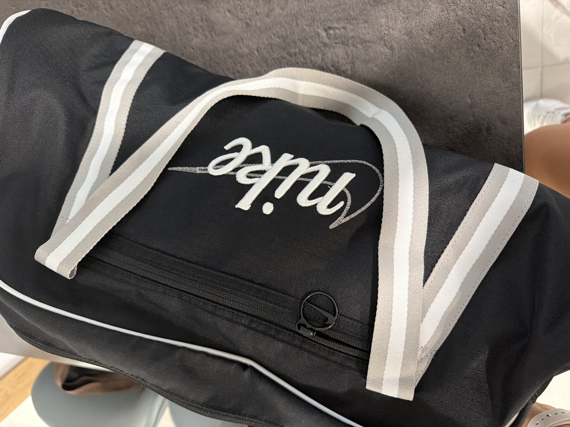 A black Nike duffel bag with white and gray striped straps is laid out on a dark gray, textured surface, likely a table. The Nike logo is embroidered in white script on the front of the bag, and a zipper with a circular pull tab featuring a Nike swoosh is visible. The image is taken from a high-angle perspective. The location of Finestrat, Spain, is not depicted in the image.