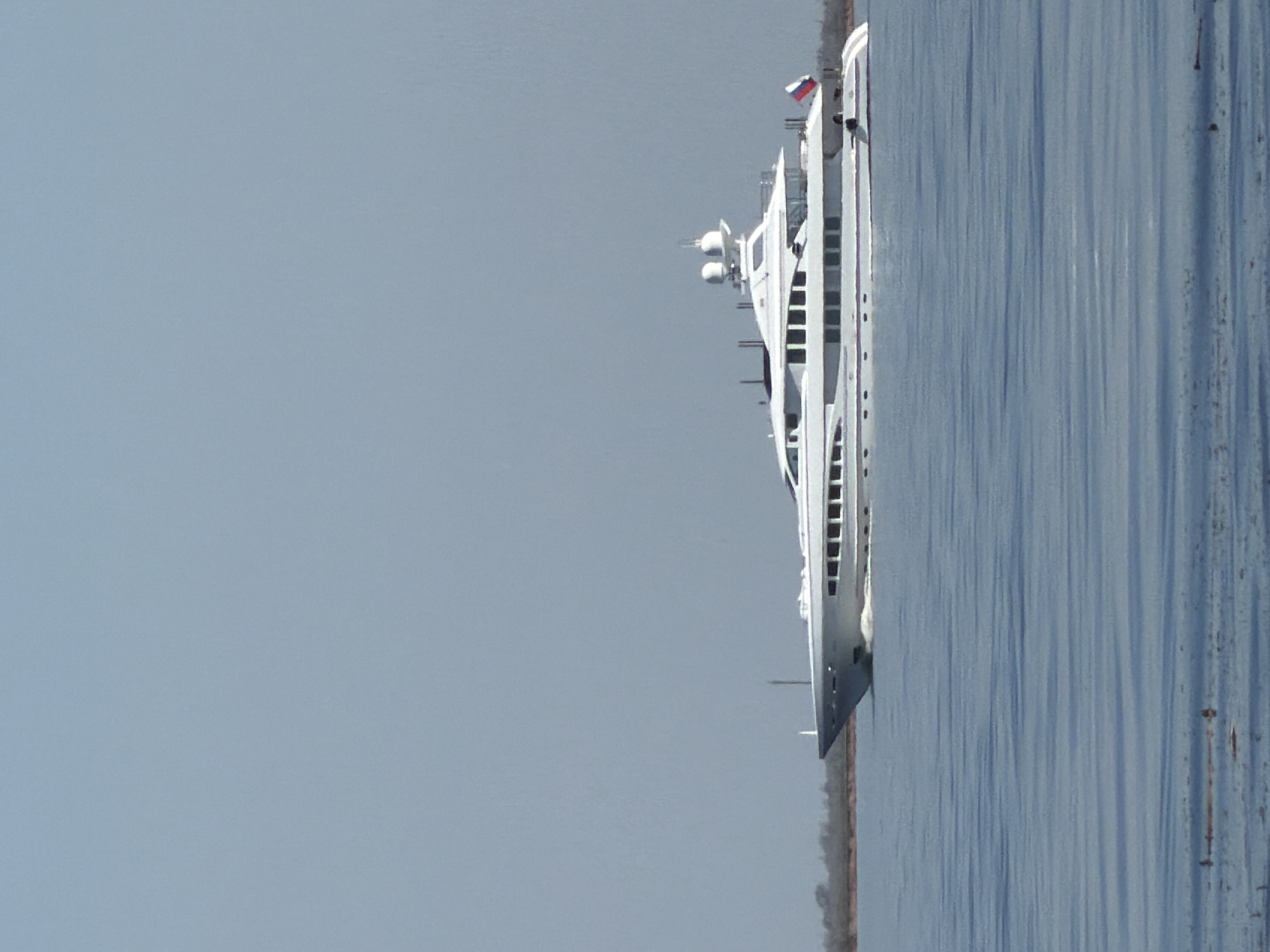 A white ship with a Russian flag is traveling along a gray body of water. A thin strip of land is seen on the bottom right. Most of the image is a cloudy light blue sky. The location appears to be Kamyshin, Russia.