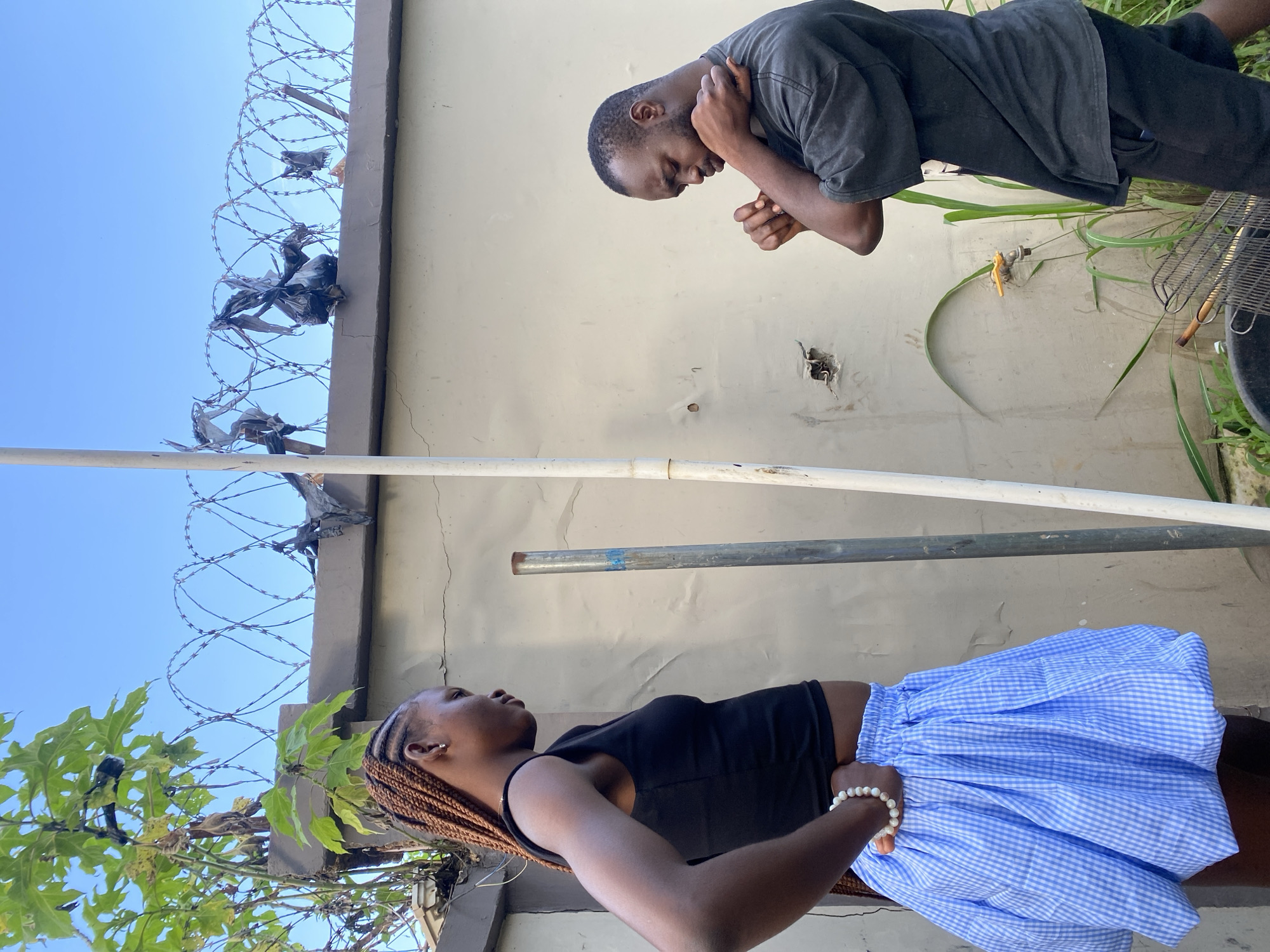 Two individuals are standing in front of a light-colored wall in an outdoor setting, under a clear blue sky. A metal fence with barbed wire is visible at the top of the wall. On the left, a person with braided hair is wearing a black tank top and a blue and white checkered skirt, with their hand on their hip. On the right, a person in a dark t-shirt and black shorts is looking down with their arms crossed over their chest. The ground in front of them has some greenery and what appears to be part of a barbecue grill. The scene is likely in a residential area.