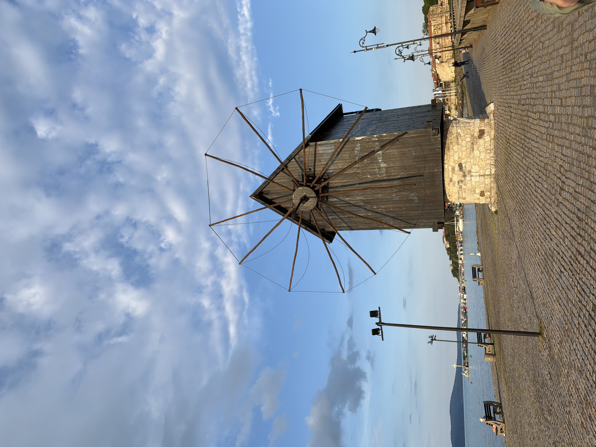 A low-angle view of a windmill in Nessebar, Bulgaria, under a blue sky with scattered clouds. The windmill's structure is dark, likely made of wood, with a series of radiating support arms. The base is constructed of light-colored stone. A cobblestone promenade runs along the right side, featuring black lamp posts and benches. Beyond the promenade, the sea stretches towards a distant shoreline dotted with buildings. The windmill sits near the edge of the town, facing the water.