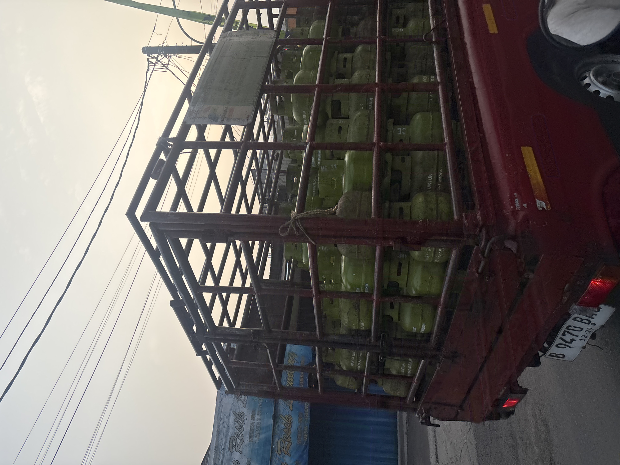 The image depicts a red pickup truck loaded with green propane gas cylinders in West Jakarta, Indonesia. The truck's bed is covered by a metal cage, and the cylinders are stacked neatly inside. Some cylinders have text printed on them, which appears to be in Indonesian. A sign is attached to the cage, likely providing information about the cargo or delivery. In the background, there is a building with a blue facade and some awnings, as well as electrical wires strung across the sky. The sky is overcast, suggesting it might be early morning or late afternoon. The license plate of the truck is visible, reading "B 9470 BAQ". The overall scene suggests a commercial delivery of gas cylinders within a city environment.