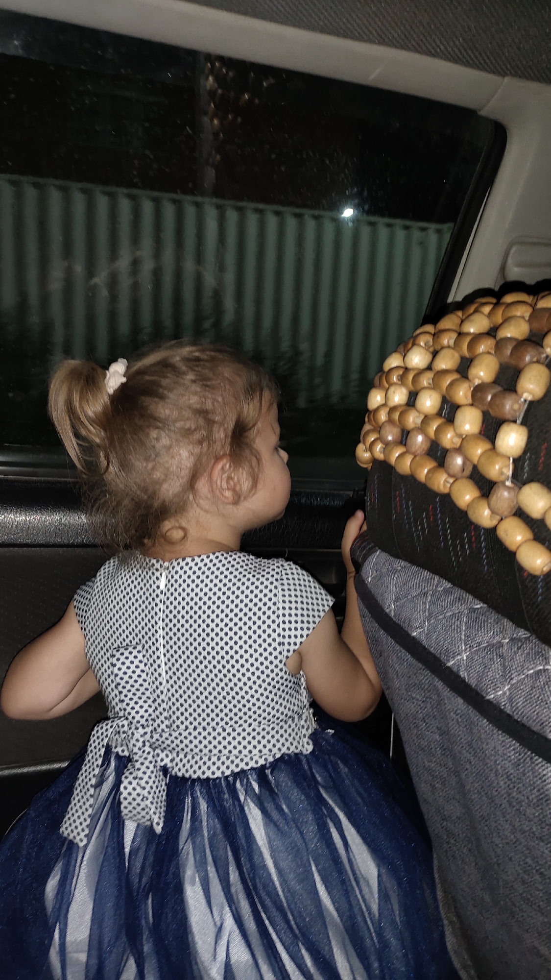 A young girl is sitting in the backseat of a vehicle, looking out the window. She has blonde hair in a ponytail and is wearing a navy blue and white polka dot dress with a sheer navy blue overlay. The girl's arm rests on the back of the driver's seat, which is covered with a beaded seat cover. The window shows a dark, outdoor scene with a corrugated metal fence. The interior of the car is dark, suggesting it is nighttime.