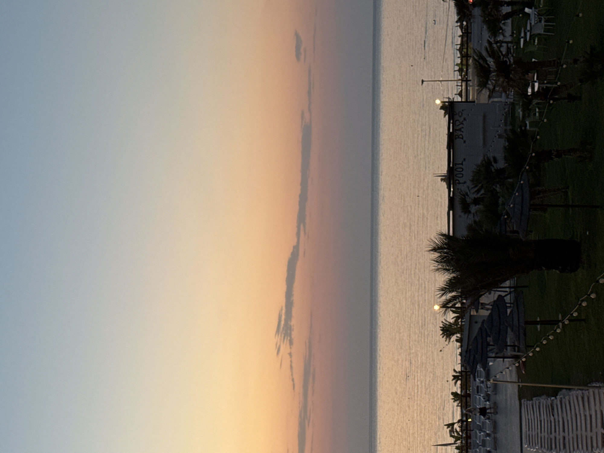 This is a photograph of a sunset scene at a beach resort in Isola delle Femmine, Italy. The sky transitions from a pale blue at the top to orange and pink hues near the horizon, with thin clouds scattered across. The sea is calm and reflects the warm colors of the sky. In the foreground, a resort area is visible with rows of lounge chairs, some with umbrellas, and palm trees. A building labeled "POOL BAR" is also present. A string of fairy lights is visible, illuminating the grass area. A person is standing in the middle distance, near the lounge chairs, and another person appears to be walking away.