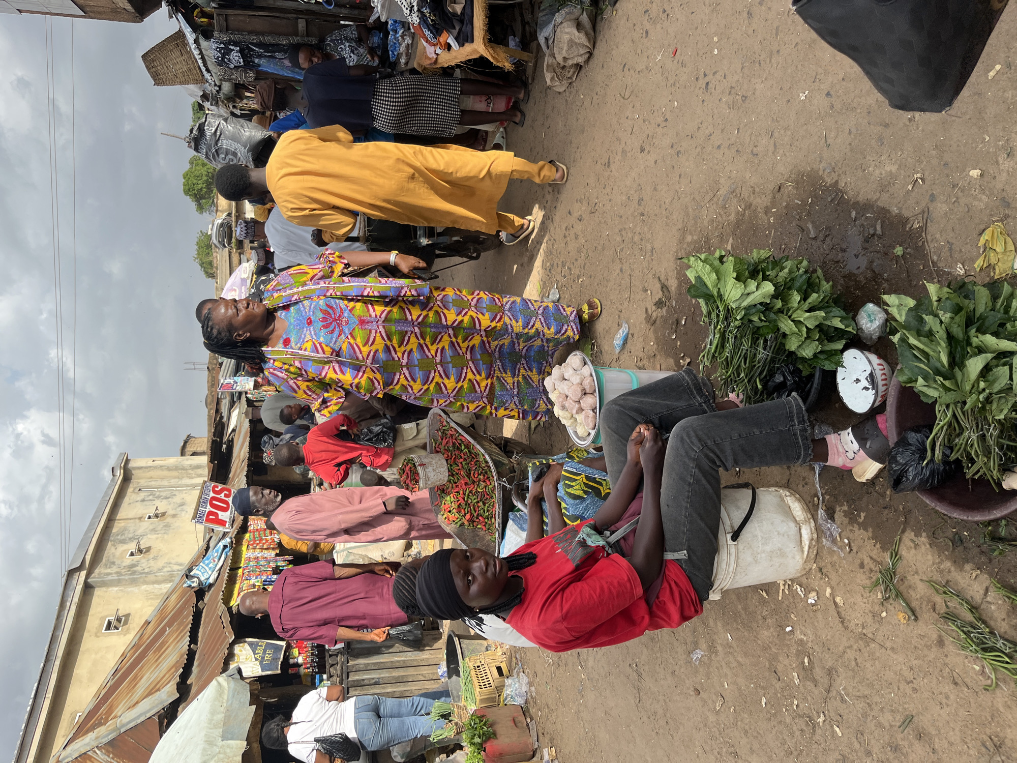 The image depicts a bustling outdoor market scene, likely in Rafin Par, Nigeria, under a cloudy sky. In the foreground, a young woman wearing a red t-shirt, dark jeans, and a black headwrap sits on a white bucket. Her braided hair is visible. In front of her, to her left, is a pile of green leafy vegetables in a dark basin, and a white bowl with some food items. To her right, a wheelbarrow is filled with red chili peppers.
A woman in a vibrantly colored, patterned dress stands in the middle ground, looking to her left. Behind her, a man in a yellow robe walks away from the camera. Other people are scattered throughout the scene, some standing and talking, others walking.
The market stalls are constructed with corrugated metal roofs and wooden structures. Signs are visible, including one that reads "POS". Merchandise, including what appears to be food and clothing, is displayed on tables and in baskets. The ground is a mixture of dirt and some wet patches, with scattered debris. The overall impression is one of a lively and active marketplace.