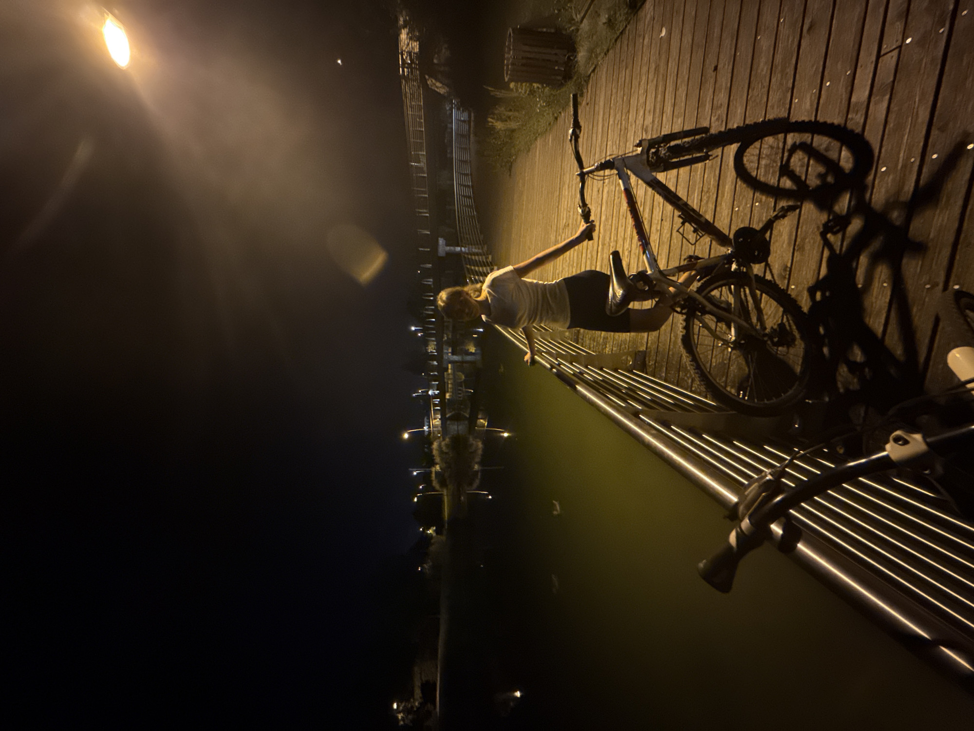 A person stands on a wooden boardwalk next to two bicycles at night. The person, wearing a light-colored t-shirt and dark shorts, is facing away from the camera and looking out over a body of water. The boardwalk is illuminated by overhead streetlights, casting long shadows from the bicycles. The water reflects the lights from the streetlights and other structures in the distance. In the background, there are bridges and trees. The scene appears to be in a park or recreational area. The presence of streetlights and the style of the boardwalk suggest an urban or semi-urban environment.
The location is identified as a boardwalk along a canal in Trento, Italy, possibly near the Adige River. The lighting suggests it is evening or night. The canal is dark and appears to have some debris or vegetation floating on the surface. The boardwalk is made of wooden planks and has a metal railing. The person is standing near the railing, with one hand resting on it. The bicycles are parked on the boardwalk, with one of them partially in the foreground. The overall atmosphere is calm and serene.