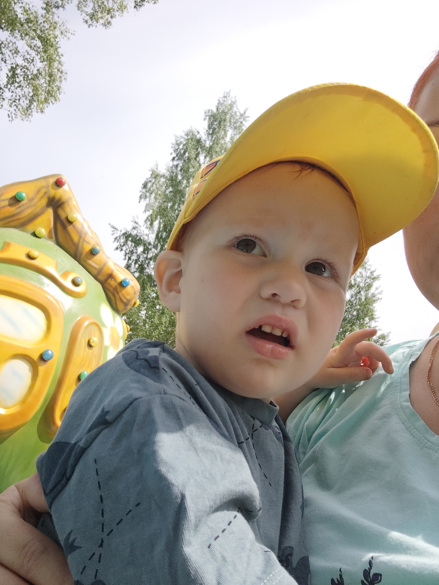 A medium, close-up shot of a young child wearing a yellow cap and a gray-blue shirt with dark stitched detailing. The child's expression shows slightly parted lips and a serious gaze upward. The photograph is taken at an upward angle, revealing elements of the surroundings. A section of a colorful, round climbing structure with yellow and green accents is visible to the left. Behind the child, there are green trees, suggesting an outdoor setting. A person, likely an adult, is holding the child. The person's shoulder and part of a light blue shirt are visible, with a gold necklace indicating a feminine appearance. Given the presence of climbing equipment and greenery, the location appears to be a park in the city of Chaikovsky, Russia.