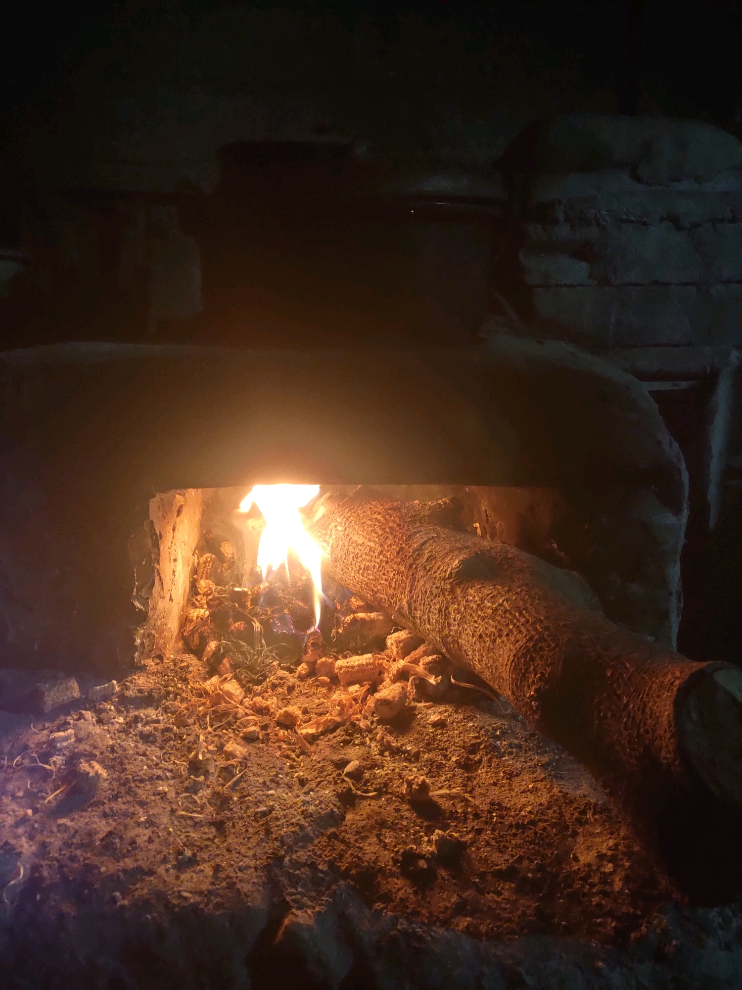 In the city of Plemahan, Indonesia, an image captures a rustic scene depicting a wood-fired oven or hearth. The opening of the oven is illuminated by the bright orange and yellow flames of a fire. Inside, remnants of burnt wood and organic material are visible amidst the glowing embers. A thick log, partially charred, rests on the edge of the oven, its rough texture highlighted by the warm light. The surrounding area appears to be constructed from rough, uneven stone or earth, suggesting a traditional or makeshift cooking setup. The overall atmosphere is dark and warm, emphasizing the primal nature of fire and its use for cooking or heating. There are no people or animals present in the frame.