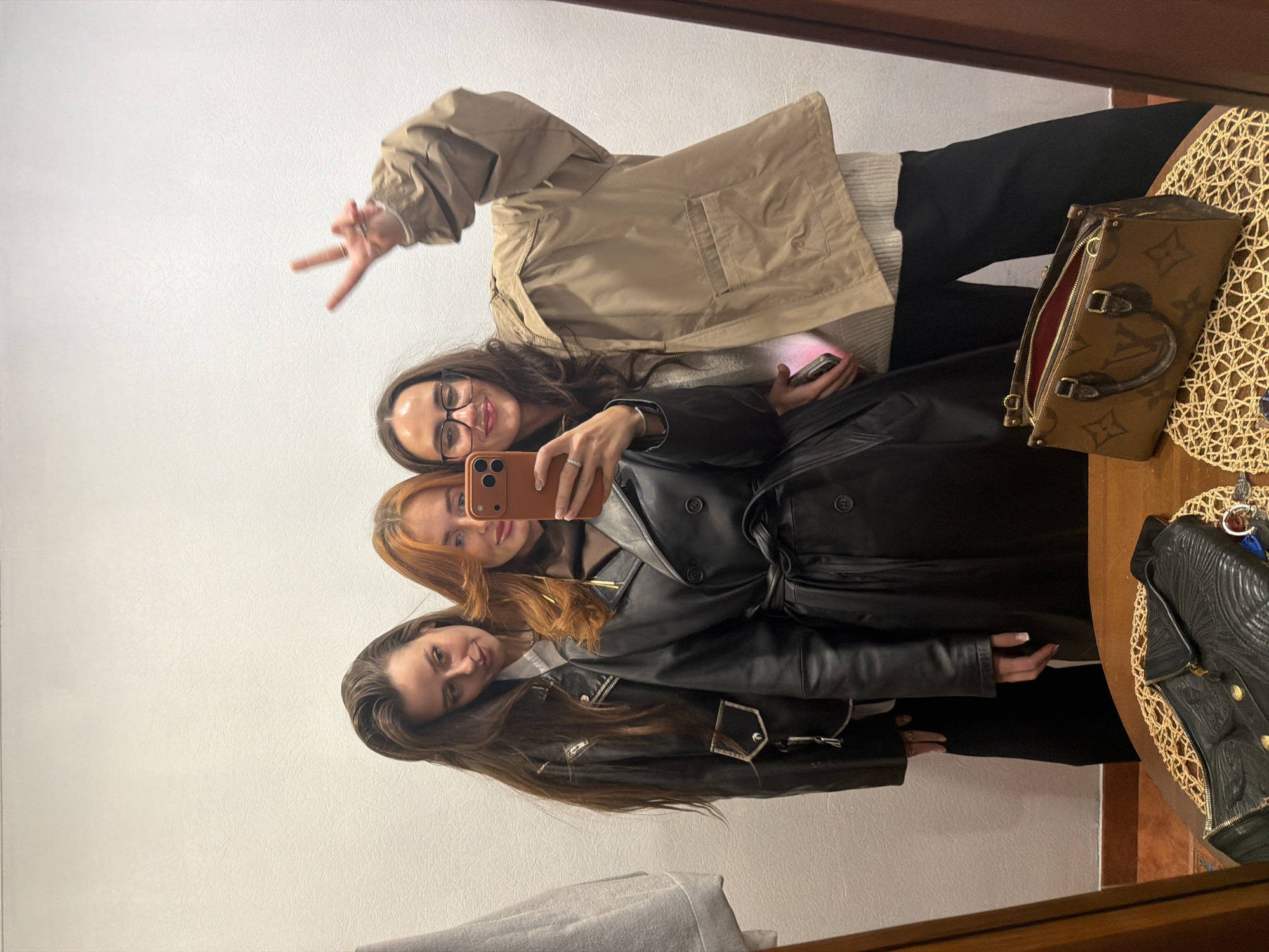 A selfie taken in a mirror shows three women in an indoor environment. The first woman, with long brown hair and glasses, is wearing a beige jacket and making a peace sign with her fingers. She is holding a phone, possibly taking the selfie. The second woman has red hair and is wearing a black leather trench coat, while the third woman has long brown hair and is wearing a black leather jacket. The background is a plain white wall. To the right of the women, a brown handbag and a black jacket are visible on a table with a woven placemat.