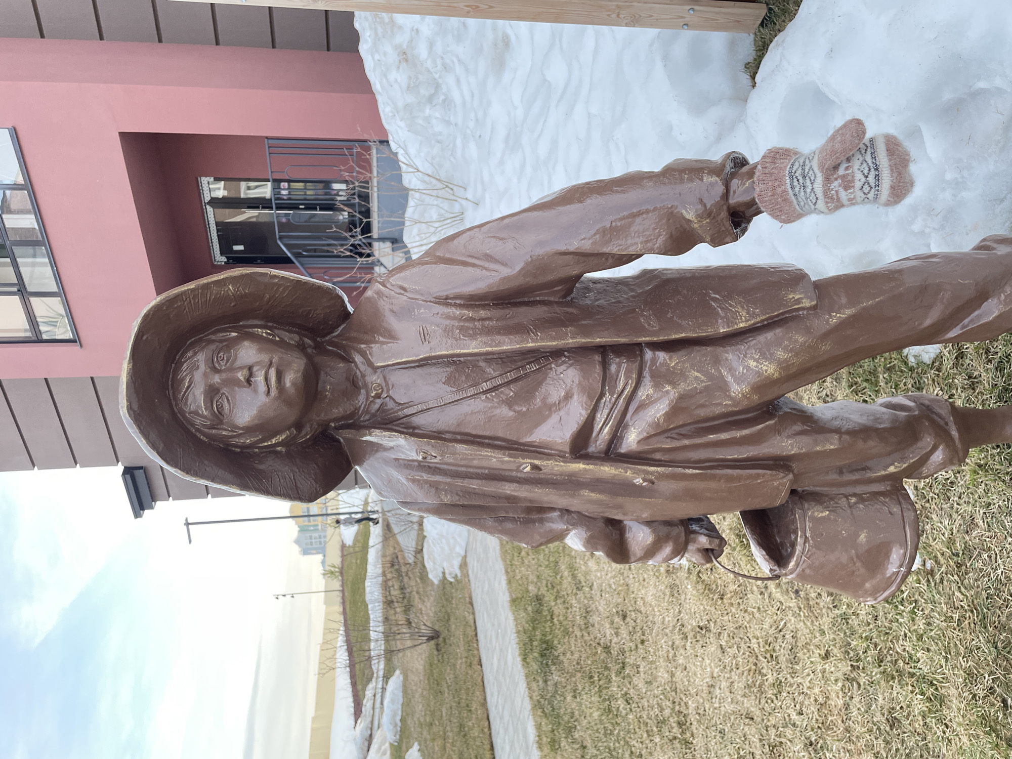 A bronze statue of a child wearing a hat and holding a bucket stands on a patch of grass. A pink knitted mitten is placed on the statue's outstretched hand. Patches of melting snow are visible around the statue and in the background. A light pink building is partially visible to the left of the statue. The background includes other buildings and a pale sky. The statue is likely in Pervomaysky, Russia, based on the prompt's context.