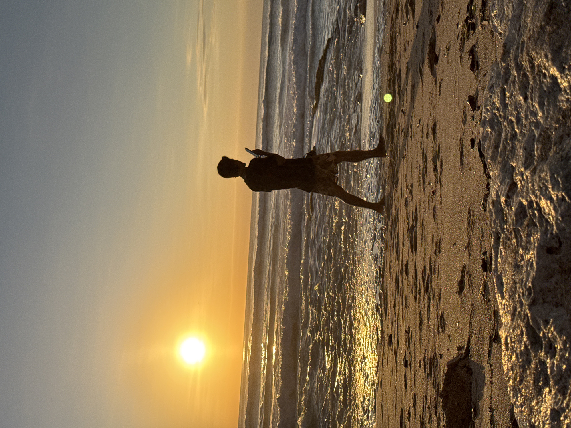The image depicts a serene beach scene at sunset in Kuta Selatan, Indonesia. The sun, a bright orb, is low on the horizon, casting a warm golden glow across the sky and illuminating the ocean with shimmering reflections. Gentle waves roll onto the sandy shore, creating a peaceful ambiance. A lone figure, silhouetted against the setting sun, walks along the beach, holding a mobile phone. The foreground of the image features textured sand with a few scattered small stones and debris. A bright green ball is visible on the sand in the mid-ground, to the right of the walking figure. The overall scene evokes a sense of tranquility and solitude.