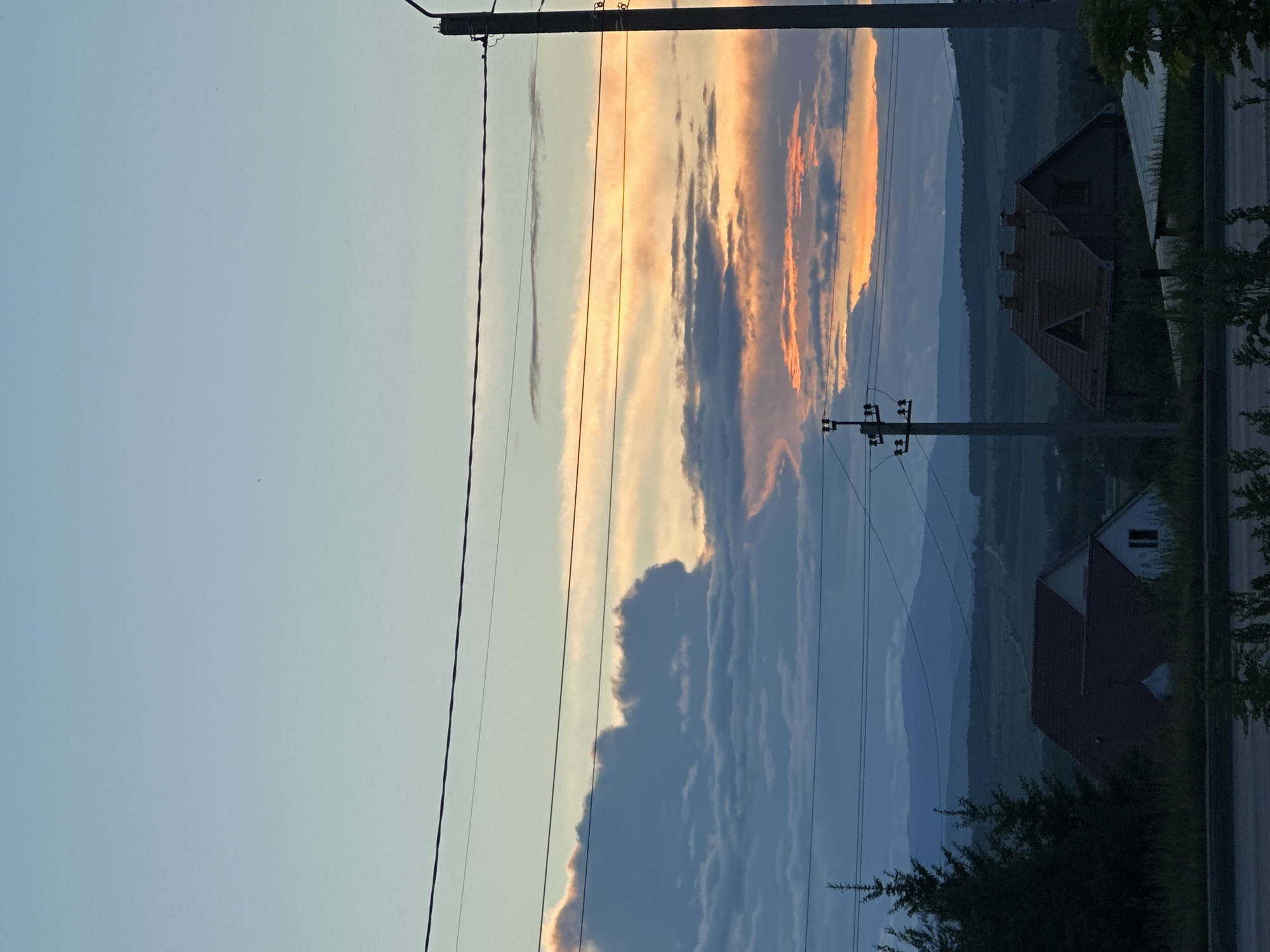 The image displays an outdoor, rural or semi-rural landscape scene captured during either sunrise or sunset, indicated by the vibrant colors in the sky.
The upper portion of the frame is dominated by a sky transitioning from a pale blue at the zenith to a dramatic palette of orange, pink, yellow, and deep gray clouds near the horizon. Large, dark cumulus clouds are visible on the left, partially backlit with warm hues, while more stratified and broken clouds with intense orange and pink illumination span the right side.
In the midground, several horizontal power lines traverse the frame, silhouetted against the brighter sky. These lines connect to a prominent, dark utility pole on the far right, extending vertically out of frame, and to a smaller utility structure with multiple cross-arms positioned slightly left of center.
Distant mountains or large hills appear as hazy, dark blue silhouettes across the horizon. Below them, a valley or cultivated landscape stretches out, showing variations in green and brown tones.
In the foreground, two distinct residential houses are visible. On the left, a white house with a dark red gable roof is partially obscured by dense, dark evergreen trees and other foliage. On the right, a darker-toned house with a red tiled roof, visible chimneys, and a single illuminated window is prominent.
The immediate foreground features a dark, horizontal strip, possibly a road, bordered by dark green bushes and grasses. No individuals are present in the scene. The overall lighting suggests the soft, indirect light of twilight. The location is Unknown, Unknown.