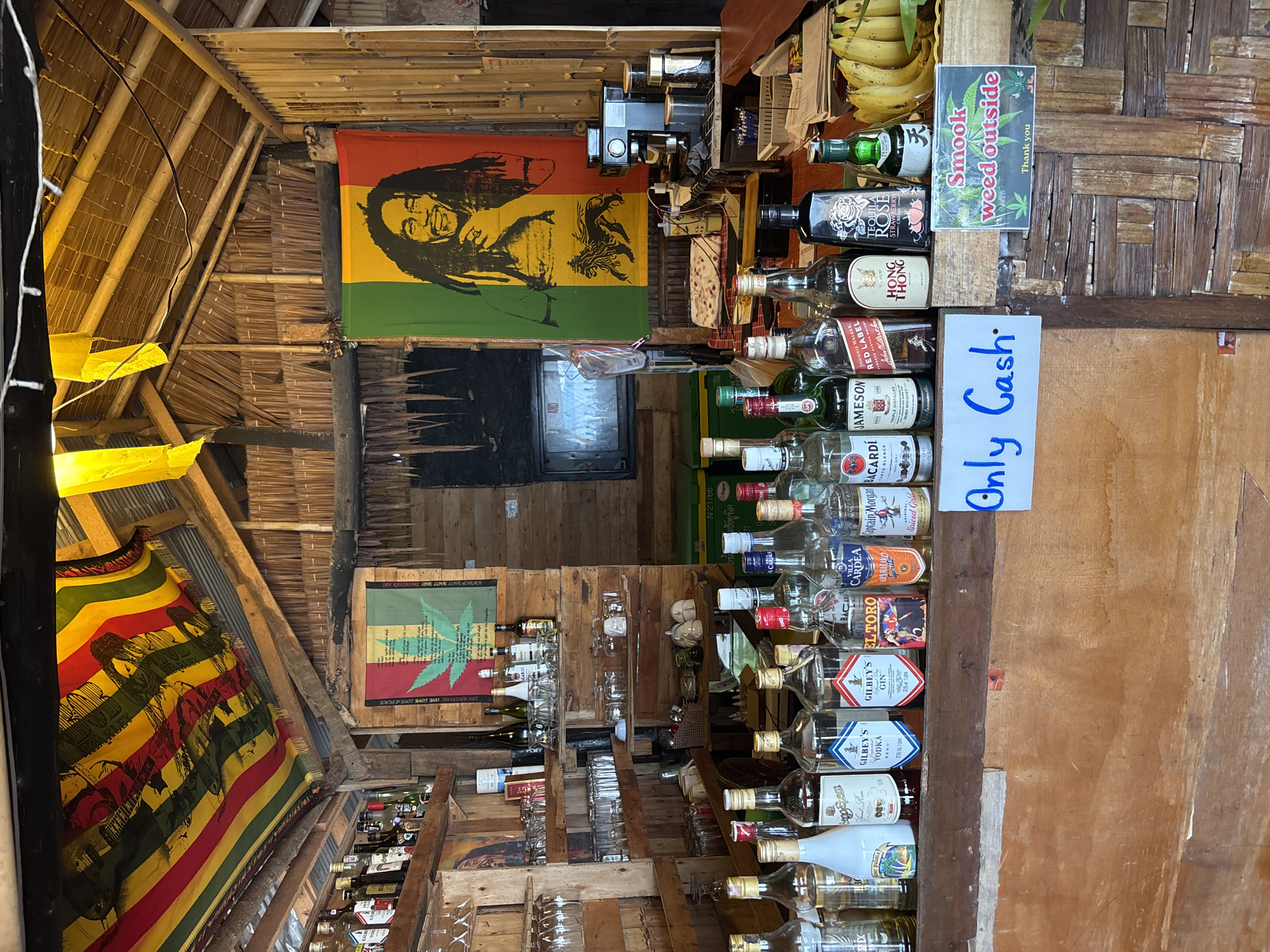 The image shows an outdoor bar with a rustic wooden construction. The bar is well-stocked with various bottles of alcohol, including gin, vodka, whiskey, and liqueurs, arranged on shelves and along the counter. In the background, there are two flags: one with a cannabis leaf design and another featuring a portrait of Bob Marley, both set against a backdrop of bamboo and thatched roofing. A coffee machine and some packaged food items are visible on the counter. In front of the bar, a sign reads "Only Cash" and another sign indicates "Smook weed outside." A bunch of bananas and some packaged goods are displayed to the right of the counter. The overall scene depicts a casual bar environment with a distinct reggae theme.