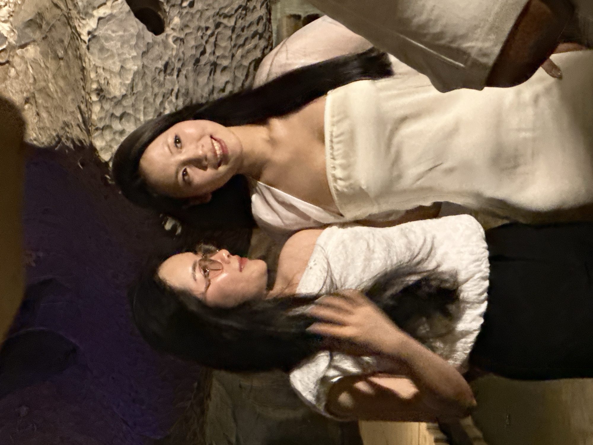 Two women are posing for a photo in what appears to be a cave. The woman on the left is wearing glasses and a white off-the-shoulder top with black pants. The woman on the right is wearing a white strapless dress and a sheer white shawl. The background is a rock formation, typical of caves. The lighting suggests the photo was taken indoors, possibly at night, with artificial lighting illuminating the scene. The location is specified as Yên Sơn Ward, Vietnam.