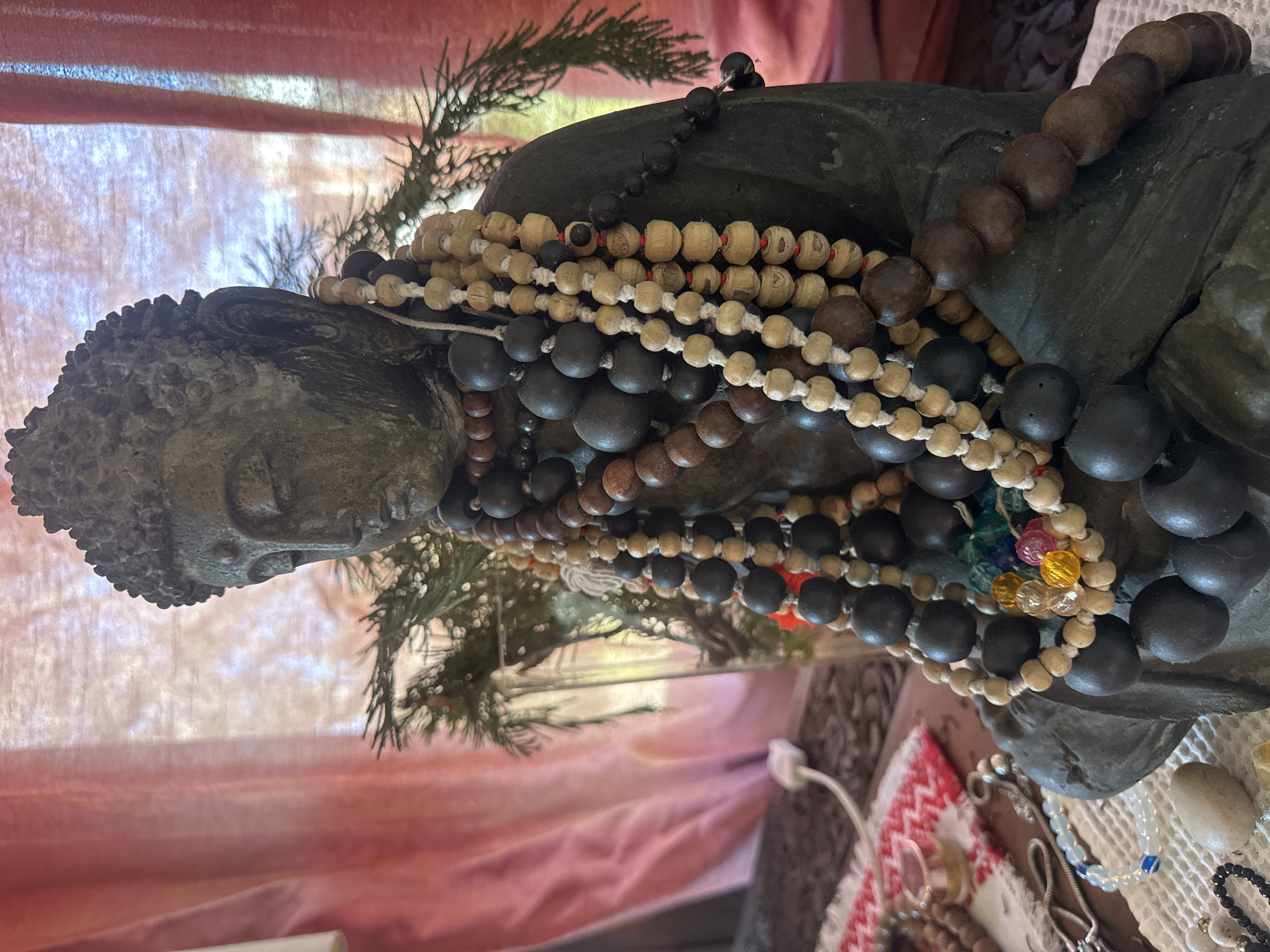 A close-up, eye-level shot focuses on a dark, stone-like statue of Buddha. The statue is adorned with numerous strands of beaded necklaces, primarily made of wood in varying shades of brown and black. Some strands also incorporate lighter-colored wooden beads and small, colorful glass beads. A sprig of evergreen branches is visible behind and to the right of the statue. The background consists of a sheer, pinkish-red curtain with soft folds, through which light filters, creating a diffused and hazy effect. The scene appears to be indoors, possibly on a table or shelf, indicated by the presence of other items like a small silver tin and what appears to be a white crocheted cloth beneath the statue and some scattered beads. The overall impression is one of a peaceful, spiritual setting. No specific location in Thora, Australia can be identified from the image.