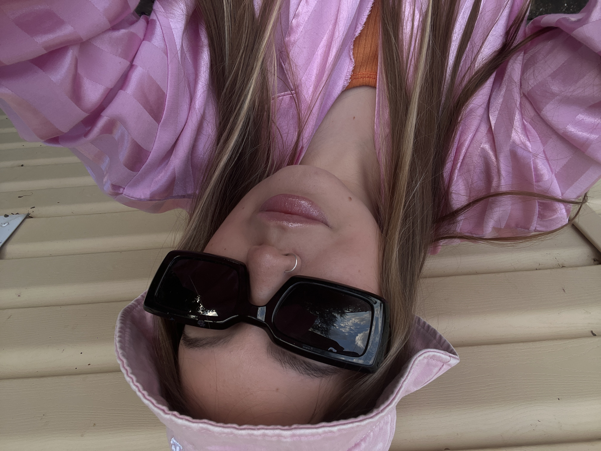 The image is an upside-down, close-up selfie of a person wearing sunglasses and a pink bucket hat. The person has blonde hair with lighter streaks and is wearing a pink, satin-like shirt with a subtle striped pattern. Underneath, a hint of an orange garment is visible. The person's lips are slightly parted, and they are wearing a silver hoop nose ring. The background appears to be a beige, horizontally-paneled surface, possibly siding on a house, suggesting an outdoor setting. The lighting and visible reflection in the sunglasses indicate it is daytime. The overall mood conveyed is casual and trendy. No text is visible in the image.