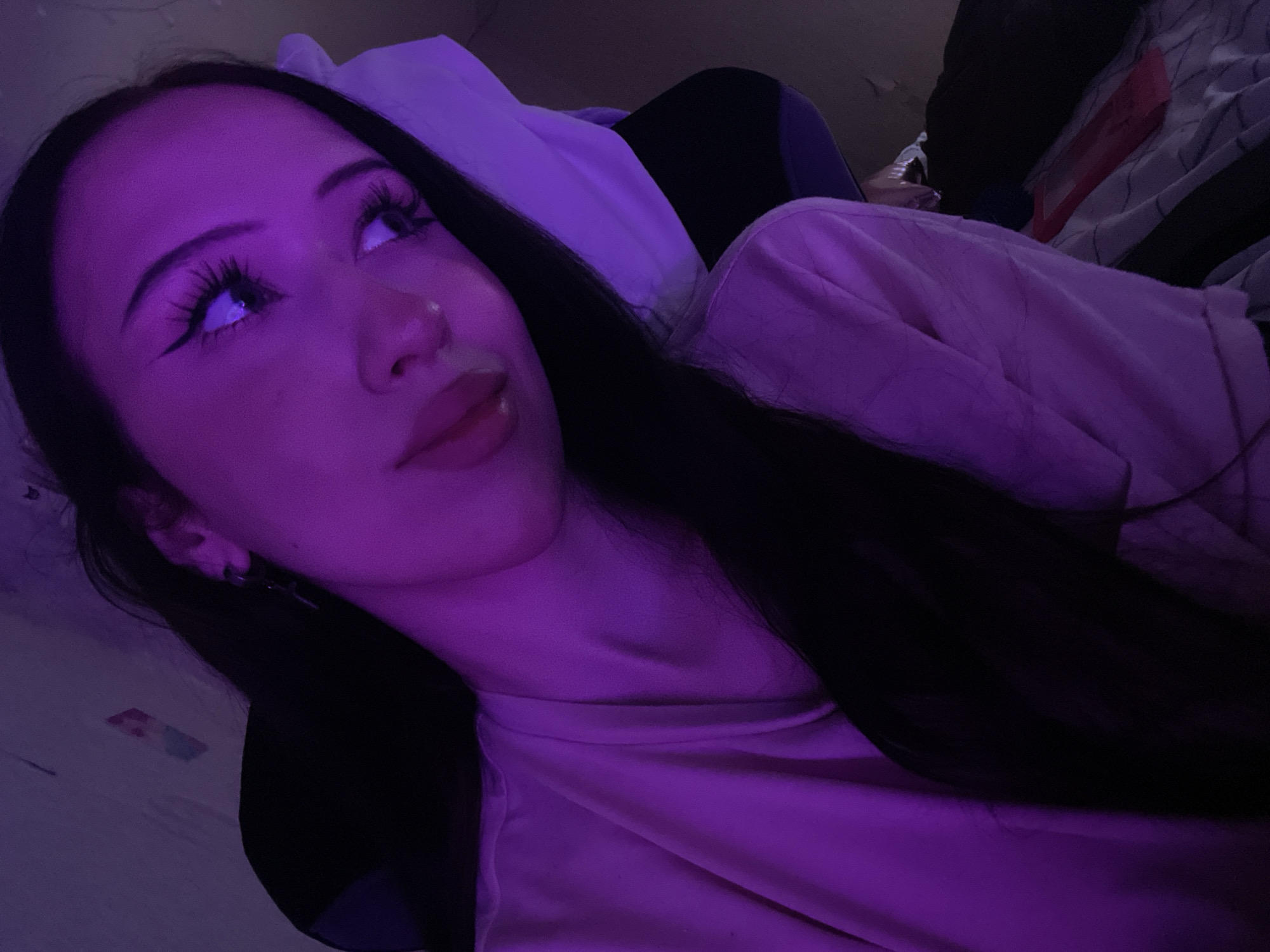 The image shows a young woman with long dark hair wearing a light-colored t-shirt. She is looking to her right, with her head tilted slightly. Her makeup includes winged eyeliner and long eyelashes. The lighting in the image is predominantly purple, casting a neon glow on her skin and surroundings. The background is indistinct, but appears to be an indoor setting with some clutter, including what looks like bedding and a pillow. There are no discernible landmarks or elements that would indicate a specific location in Votkinsk, Russia.