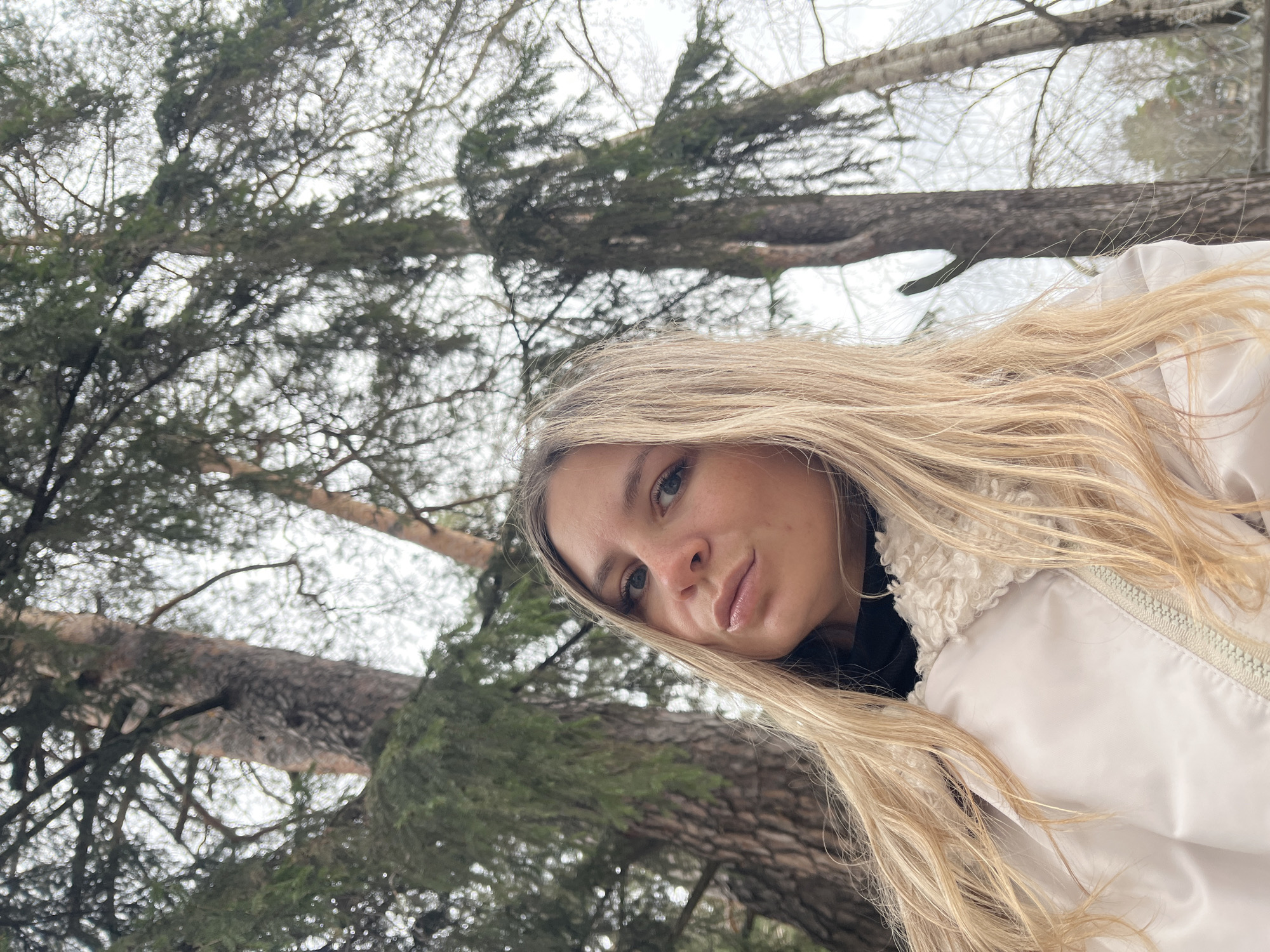 A young woman with long, blonde hair and blue eyes is captured in a close-up, tilted selfie taken outdoors in a forest, possibly near Chaikovsky, Russia. Her face, featuring a neutral to slightly contemplative expression, is positioned prominently in the foreground, looking directly at the camera. She is wearing a white puffer jacket with a textured, likely sherpa-lined, collar over a black garment.
The background reveals a dense forest canopy, viewed from a low angle looking upwards. Tall evergreen trees, with visible bark and needle-like foliage, dominate the scene. Interspersed among them are bare deciduous trees with thin branches, set against a bright, overcast sky. The soft, diffused lighting suggests a cloudy or wintery day. No discernible activity beyond taking the selfie is evident. There is no visible text.