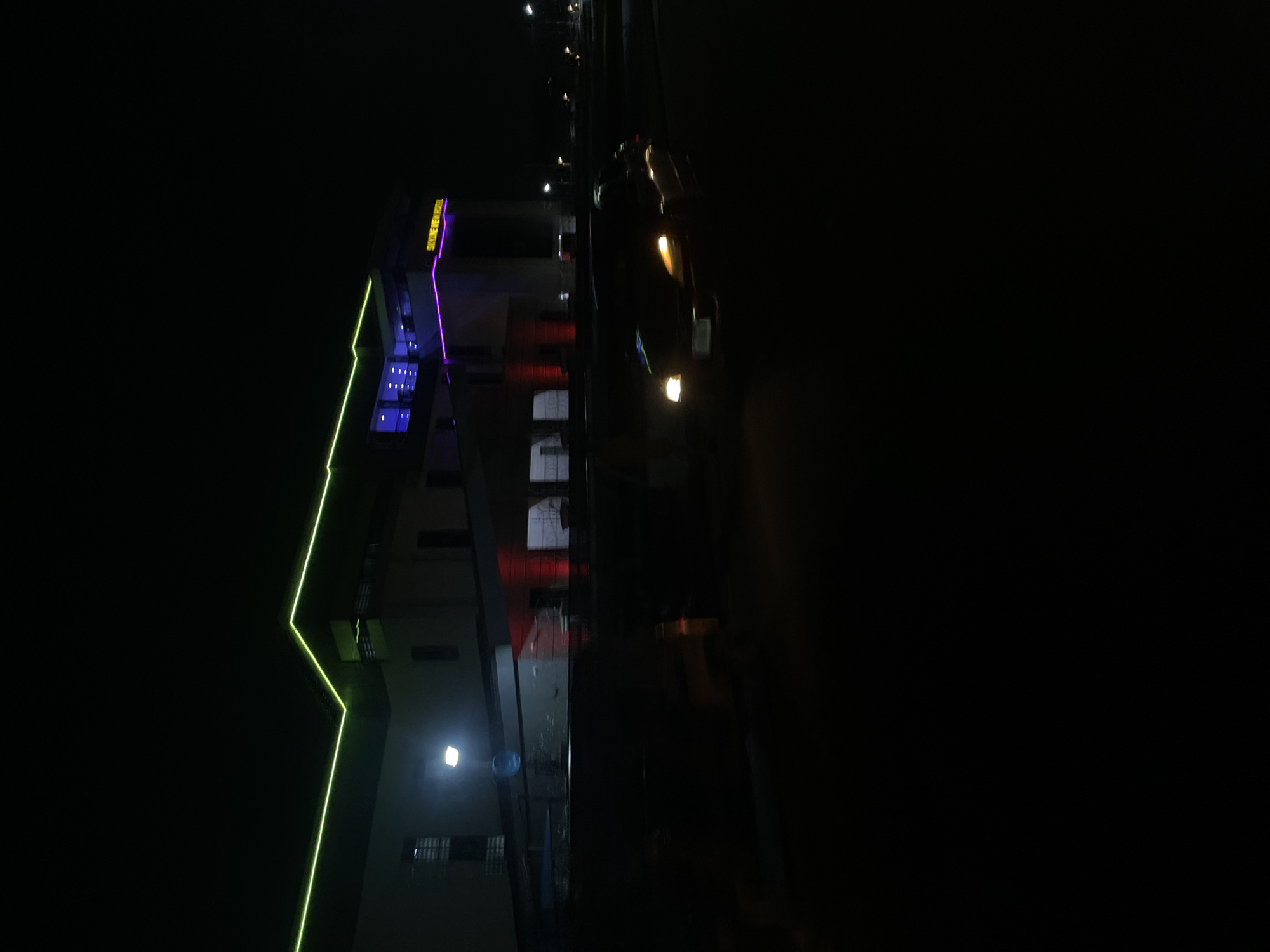 This media file presents a nighttime urban scene, likely in Abuja, Nigeria, dominated by artificial lighting. The central subject is a multi-story building, identifiable as "SKYVIEW HOTEL" from the prominently displayed yellow-amber text on its facade.
The building is strikingly illuminated with neon lights: a bright green strip traces its left vertical edge and a portion of its roofline, while the main front facade glows with vibrant purple or magenta light. Below the "SKYVIEW HOTEL" sign, a grid of small, square blue lights is visible, and further down the building, red lights add to the colorful display.
To the right of the building, a vehicle, possibly a car or truck, is seen on a road. Its bright yellow-orange headlights or fog lights cut through the darkness, though the vehicle's form and any occupants are indistinct due to motion blur or low light. The sky is completely dark, emphasizing the intense glow of the artificial lights. The scene portrays a quiet urban setting after dark, highlighting the modern, illuminated architecture of the hotel.