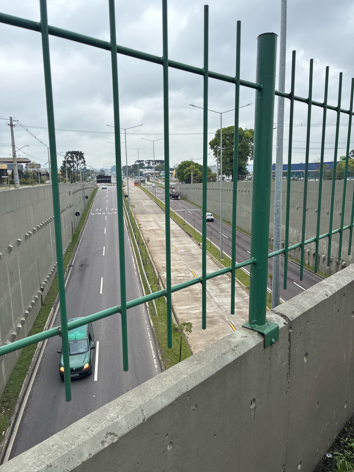 The image captures a view of the Linha Verde highway in Curitiba, Brazil, looking north. The foreground features a green metal fence and a concrete barrier. Beyond the fence, the highway stretches into the distance, characterized by multiple lanes of asphalt with white lane markings. A gray concrete wall lines the left side of the highway, with a distinct pattern of staggered indentations.
Several vehicles are visible on the highway. A dark green small van or hatchback is in the left lane, moving north. Further up the road, a white passenger car is also traveling north. In the distance, a truck is visible, as are other vehicles that are not clearly discernible.
Streetlights are positioned along the highway, and utility poles with wires are visible on the left side, indicating the urban or suburban setting. Trees and some buildings are present on the sides of the highway, suggesting an area with some development. The sky is overcast, contributing to a diffused, slightly dim lighting condition. The overall scene depicts a section of a major road in Curitiba under a cloudy sky.