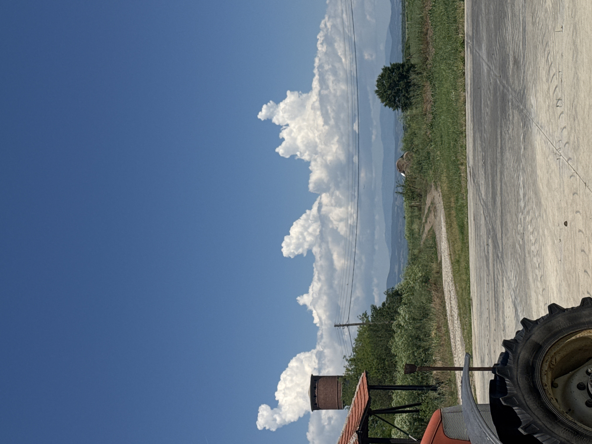 The image shows a daytime scene in Sarnevo, Bulgaria, featuring a partly cloudy blue sky and an outdoor environment. A tractor is partially visible in the foreground, with a focus on a tire. The landscape includes fields, vegetation, and a rural road. Power lines are visible against the sky and clouds. Mountains can be seen in the distance.
